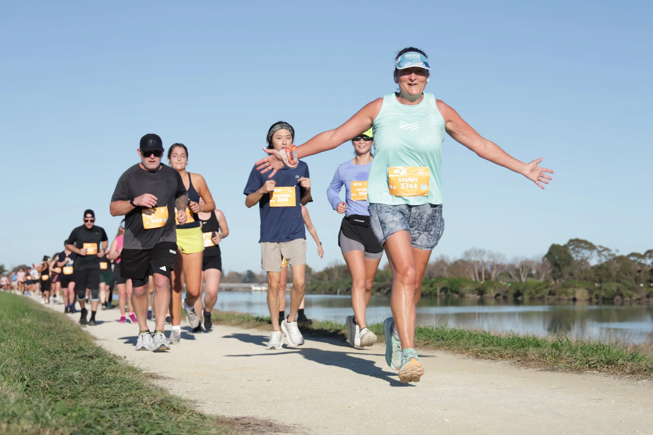 A woman with a blue visor and blue athletic outfit leading a group of diverse runners on a trail beside a body of water under a clear blue sky.