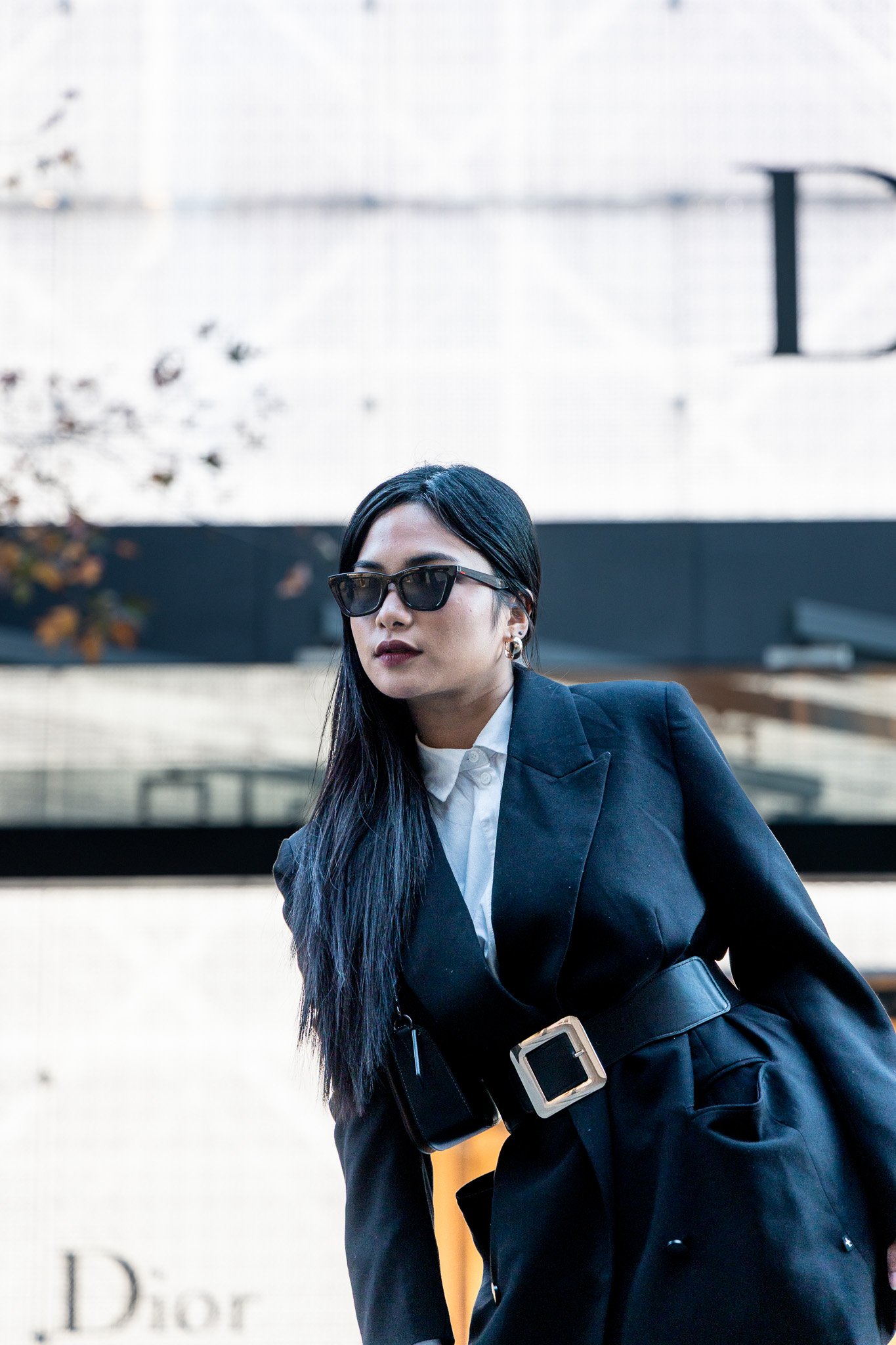 A woman wearing black sunglasses, a dark blazer, and a white shirt, with long black hair and earrings, standing outdoors in front of a white wall and trees.