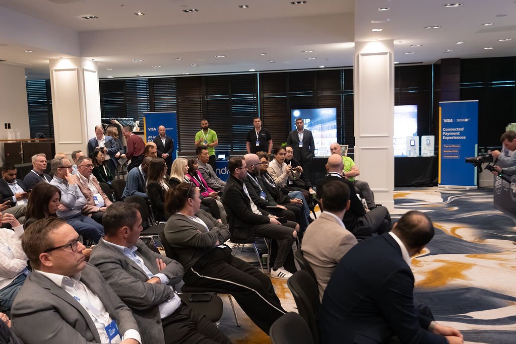 Audience attending a conference or seminar in a modern hotel conference room, with some people taking notes and a videographer filming.