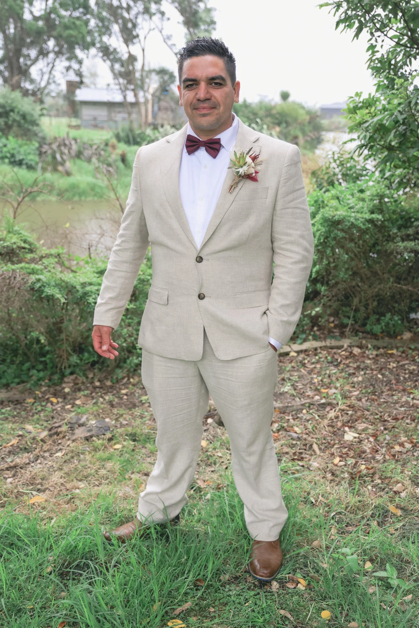 A man wearing a beige suit with a white shirt, maroon bow tie, and brown shoes, standing outdoors near a pond and trees.
