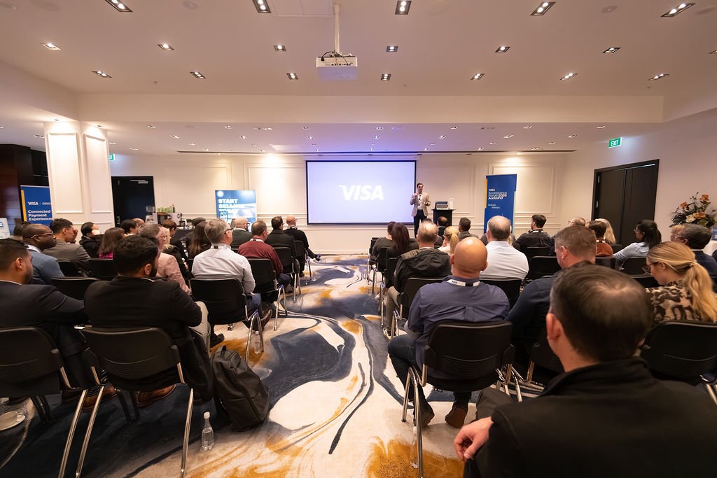 A conference room filled with attendees listening to a presenter. The screen displays the VISA logo. People are seated in rows, facing the presenter at the front.