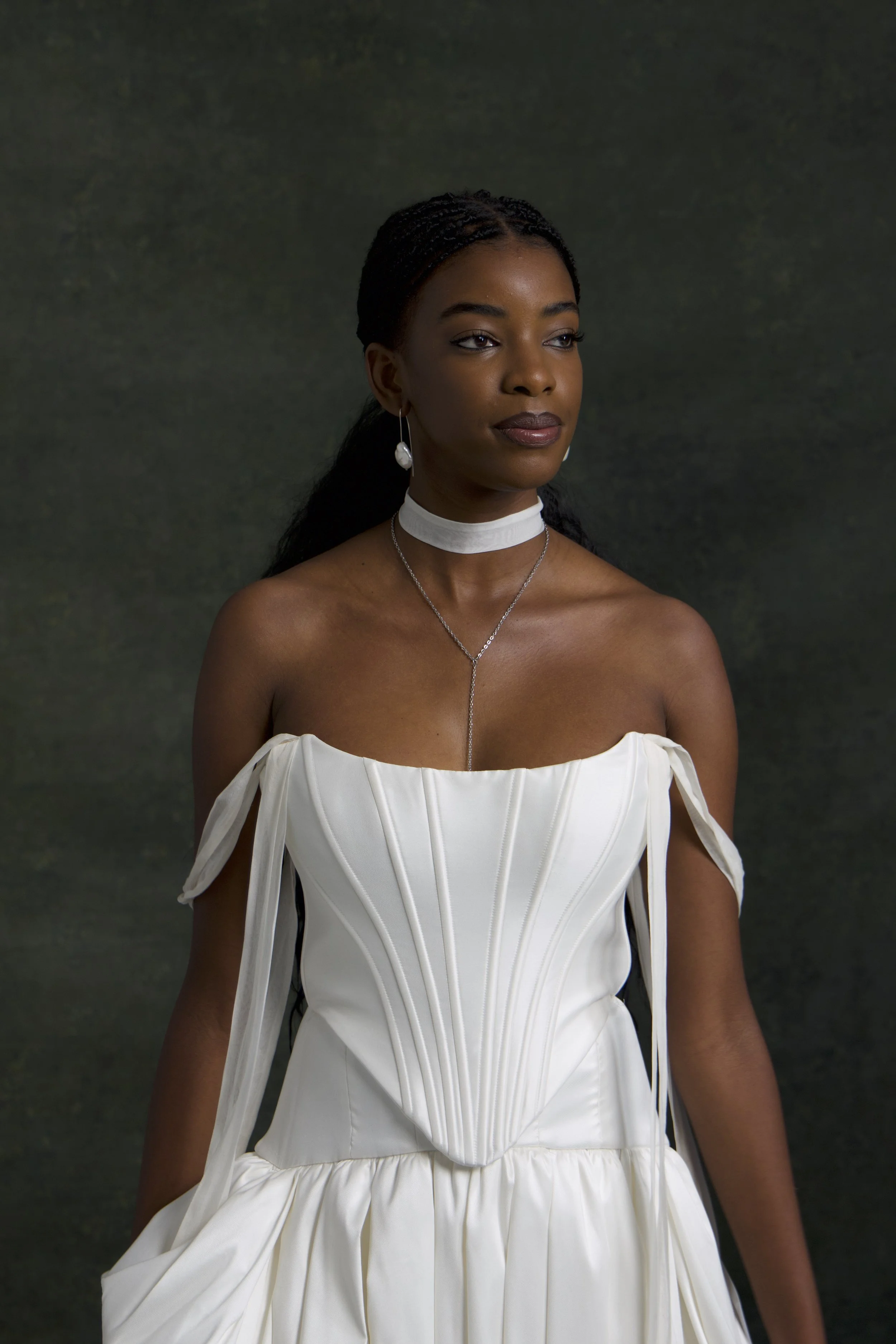 A woman with dark skin, braided hair, and pearl drop earrings wearing a white strapless dress with ruffle details and a choker necklace, standing against a dark, muted background.