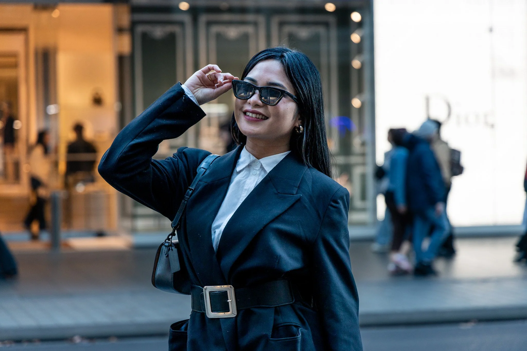 A woman with black hair, wearing a dark blazer, white shirt, and sunglasses, smiling while adjusting her glasses on a city street.
