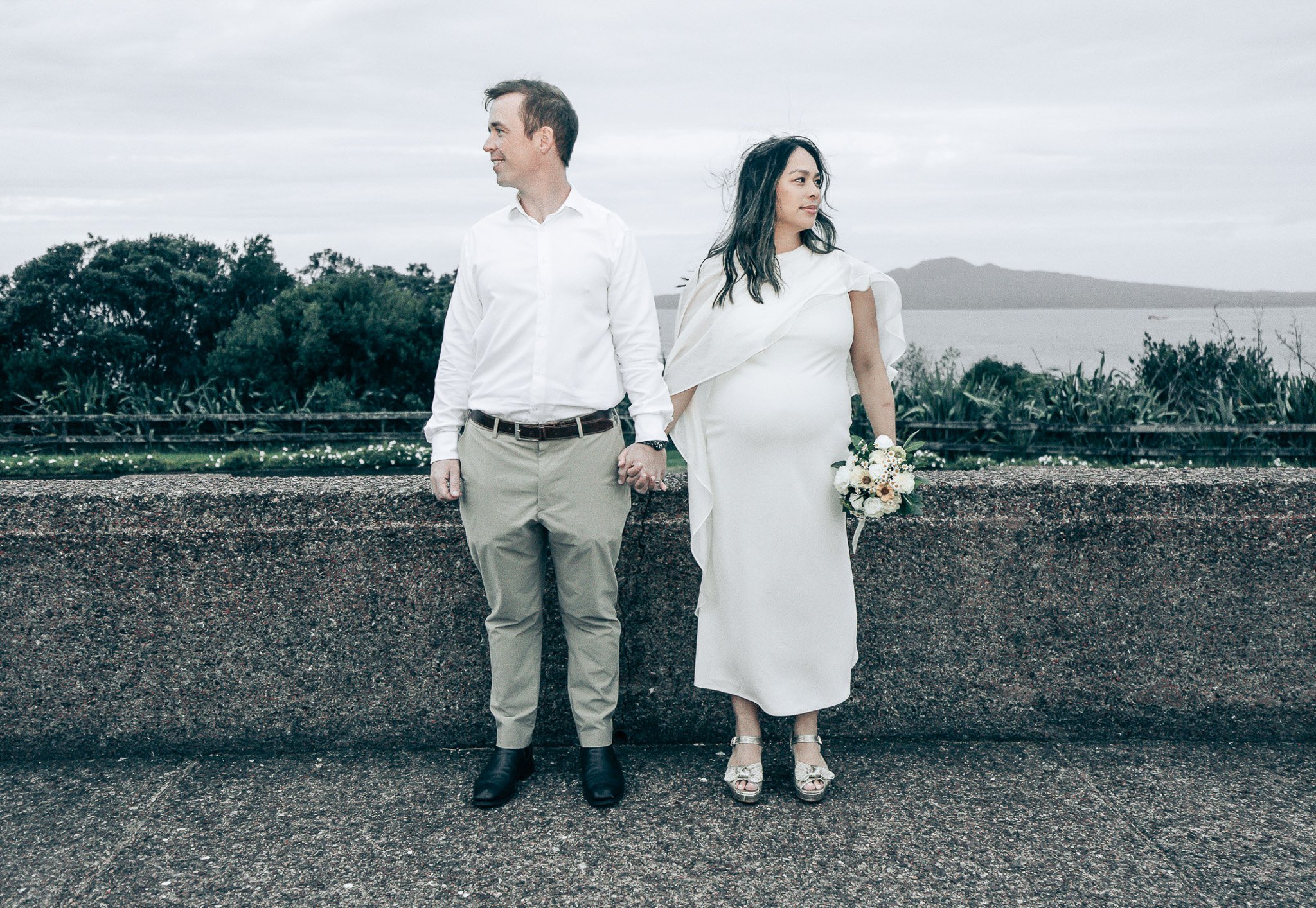 A man and woman holding hands, standing outdoors by a low stone wall with a scenic landscape in the background, the man wearing a white shirt and beige pants, the woman in a white dress holding a bouquet, overcast sky.