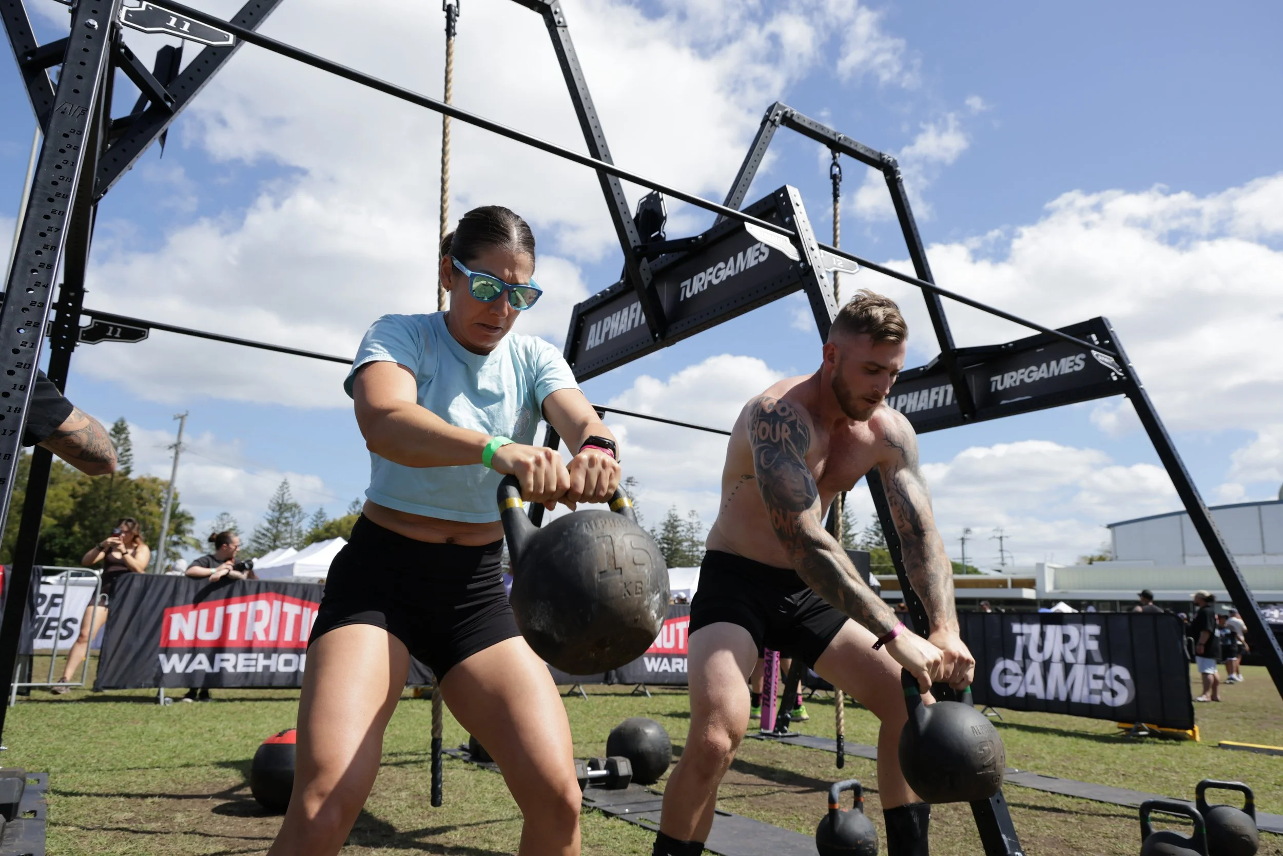 Two individuals participating in an outdoor strongman competition, lifting large black kettlebells with a grassy field, tents, and a blue sky with clouds in the background.