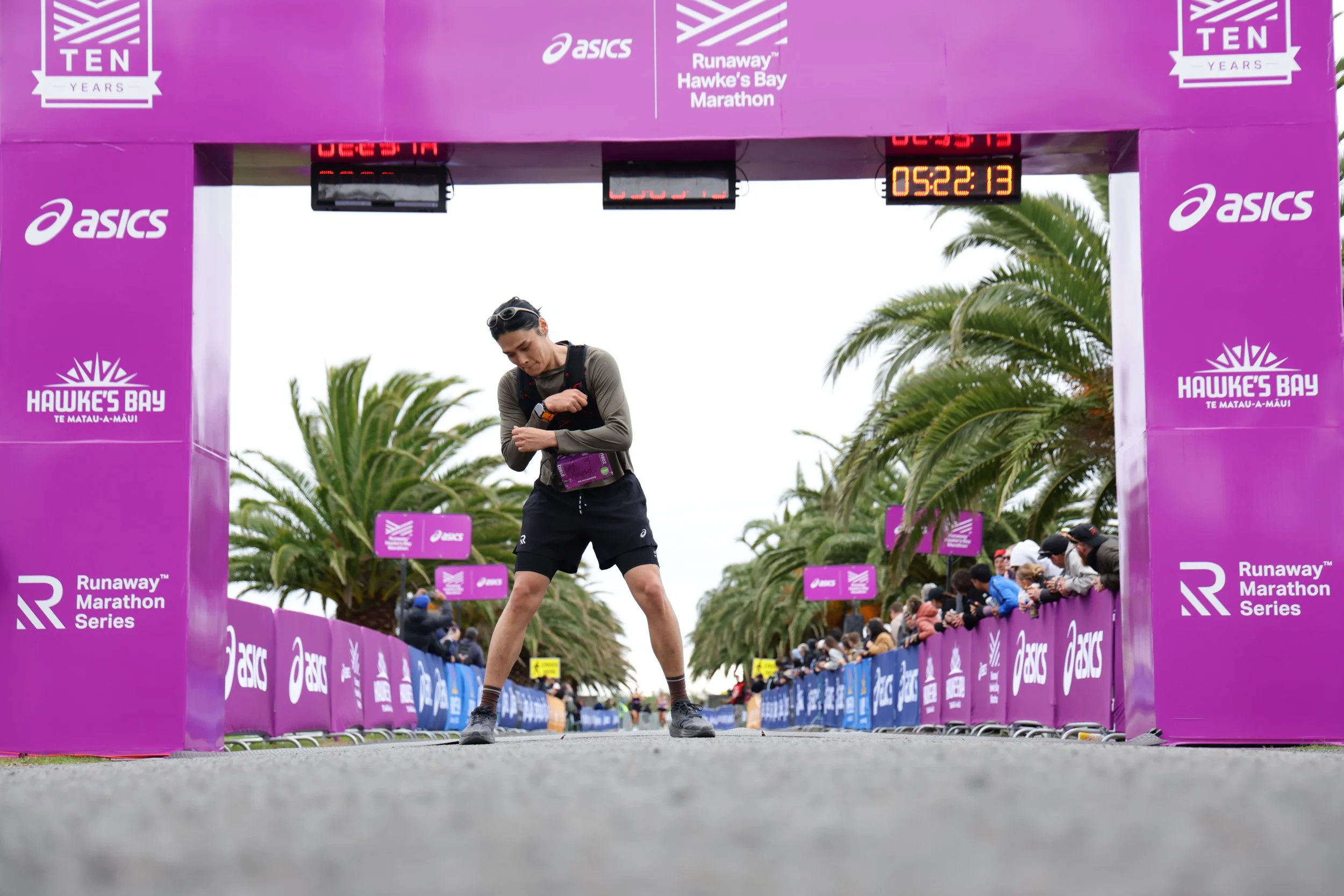 A runner crossing the finish line at the Hawke's Bay Marathon, with a purple event arch and palm trees in the background, and spectators on the sides.