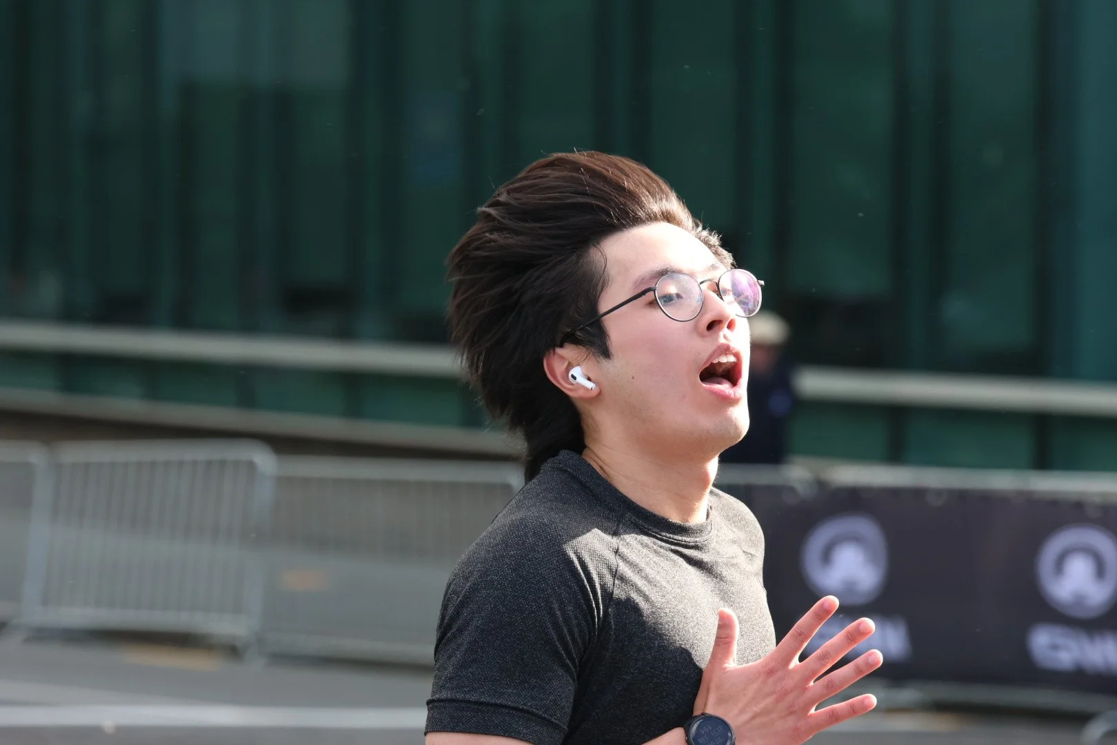 A young man with glasses running outdoors, wearing wireless earbuds, a black shirt, with a building in the background.