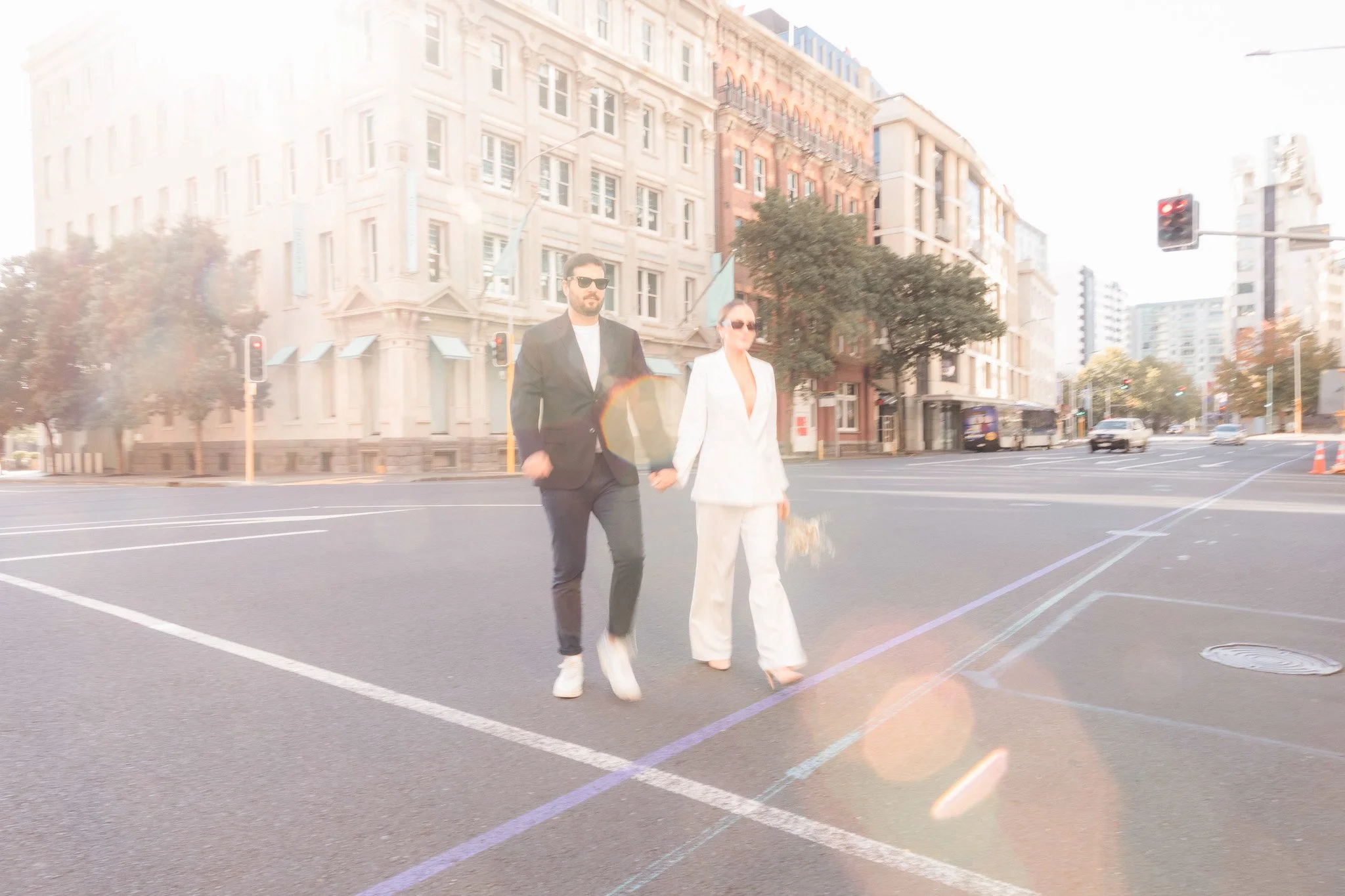 A man and woman walking hand in hand across a city street on a sunny day. The man is wearing a black suit and sunglasses, and the woman is wearing a white suit and sunglasses. The background features tall buildings, traffic lights, and moving cars.