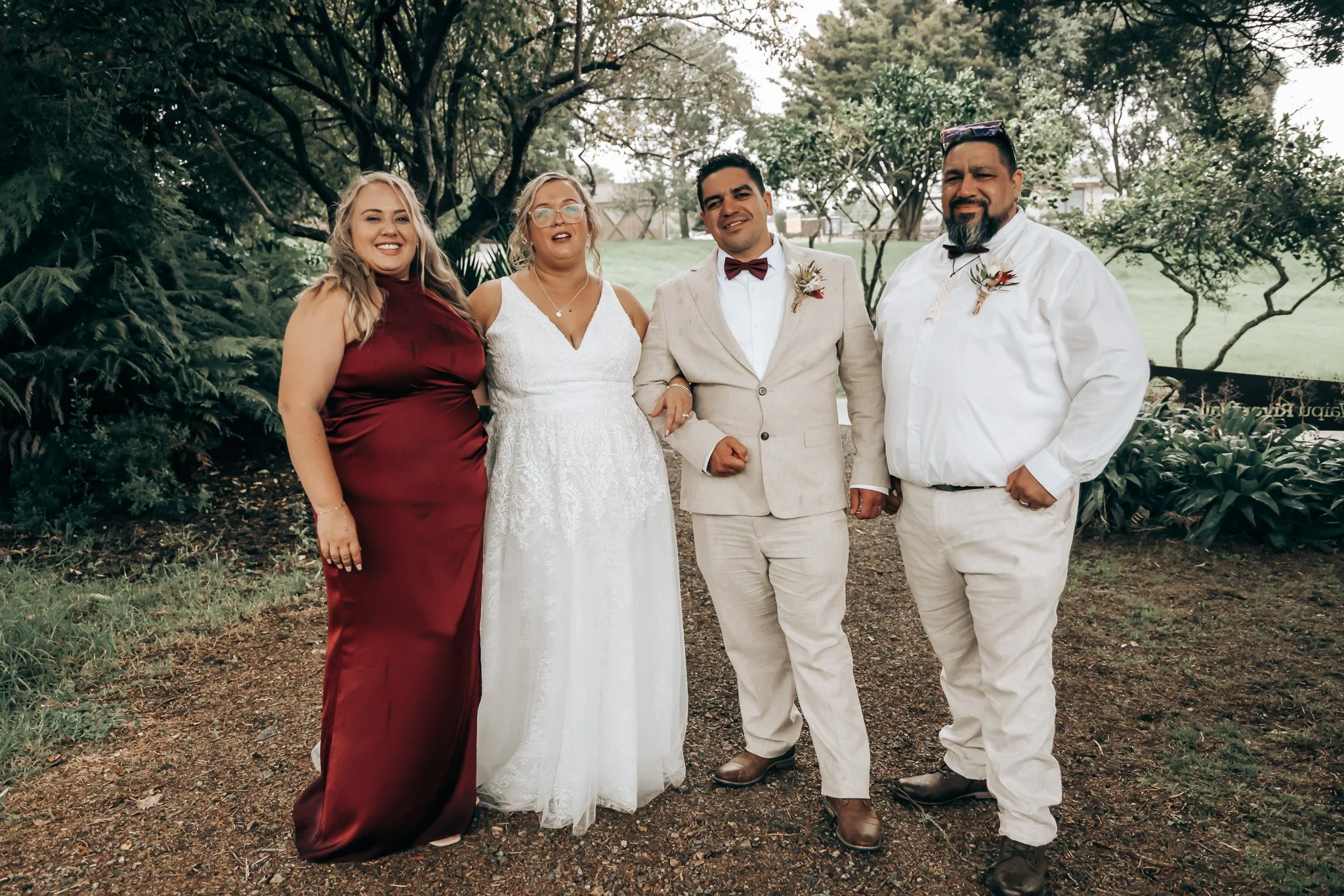 A group of five people standing outdoors on a path surrounded by trees and greenery, smiling and dressed in formal attire for a wedding.