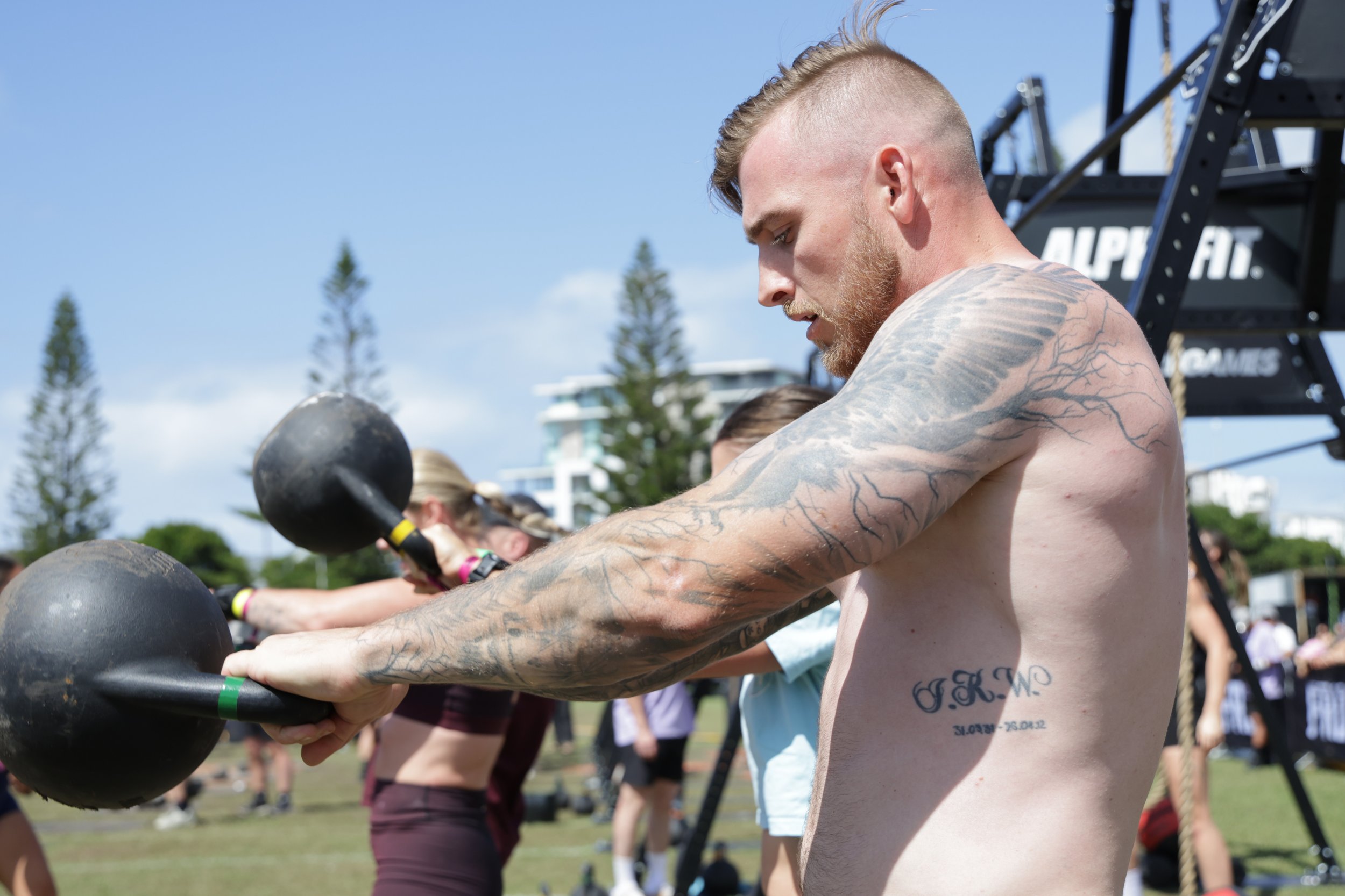 A shirtless man with tattoos on his arm and chest, working out with a large black kettlebell during an outdoor fitness event, surrounded by other participants and fitness equipment, with trees and a building in the background on a sunny day.