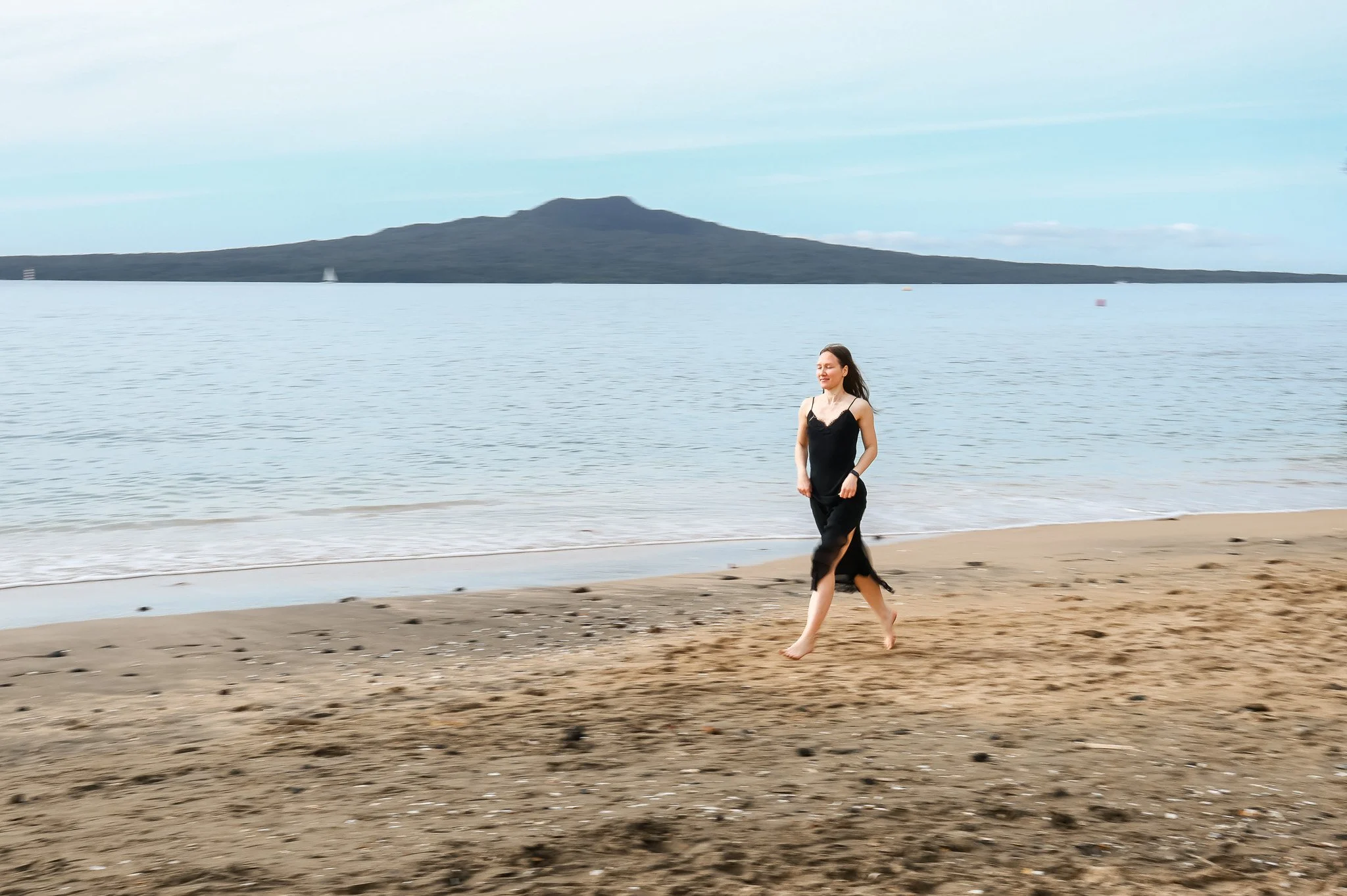 Woman in a black dress walking on a sandy beach near the water with a mountain and sailboats in the background.