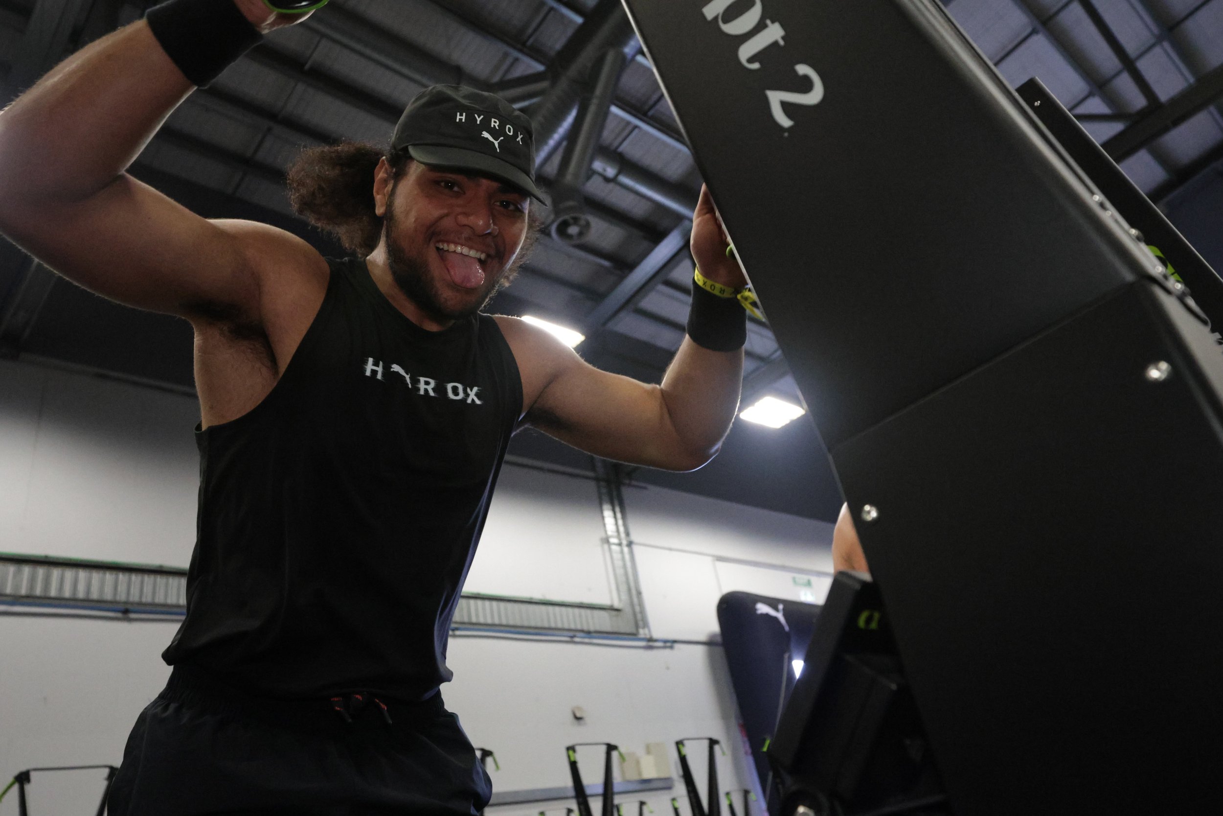A young man wearing a black tank top and a black cap is smiling with his tongue out while working out in a gym. He is lifting a weight with both hands and appears to be in a cheerful mood.