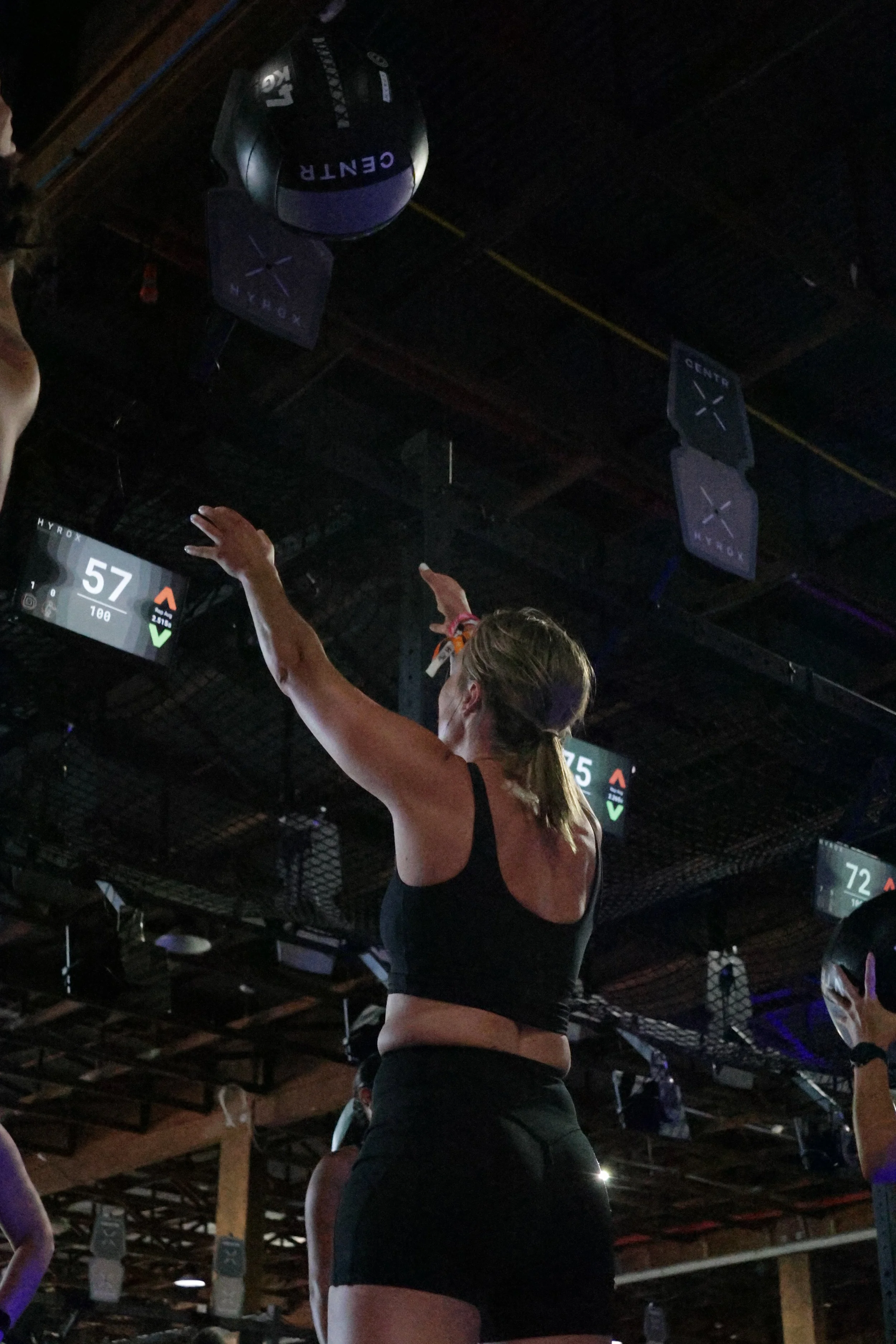 A woman in workout attire with braided hair participating in a fitness class, reaching her hands upward, in a dimly lit indoor gym with hanging signs and a screen showing the numbers 57 and 72.