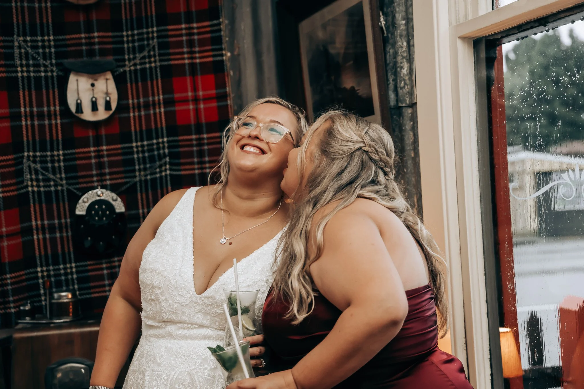 Two women with blonde hair sharing a kiss at a celebration or wedding, one in a white dress and the other in a burgundy dress, inside a decorated venue with tartan fabric on the wall and a window with rain outside.