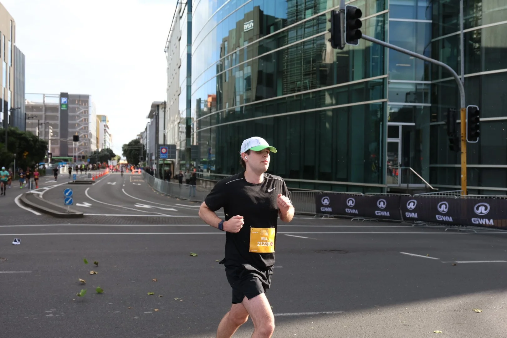 A person in running attire, including a black shirt, black shorts, a white and green cap, and a race bib with the number 21239, running in an urban street with modern glass buildings and traffic lights.