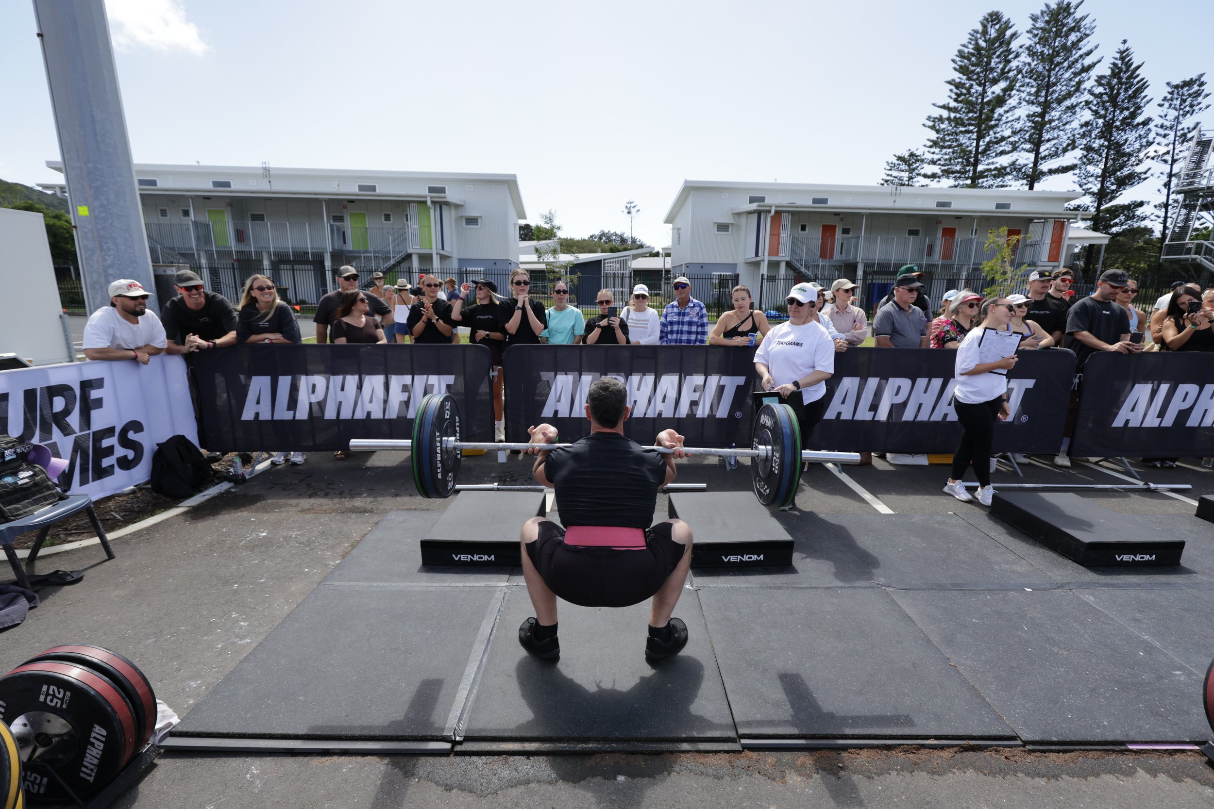 A person performing a deadlift in front of a crowd at an outdoor fitness event. The audience is behind barriers, watching and taking photos. There are buildings and trees in the background.