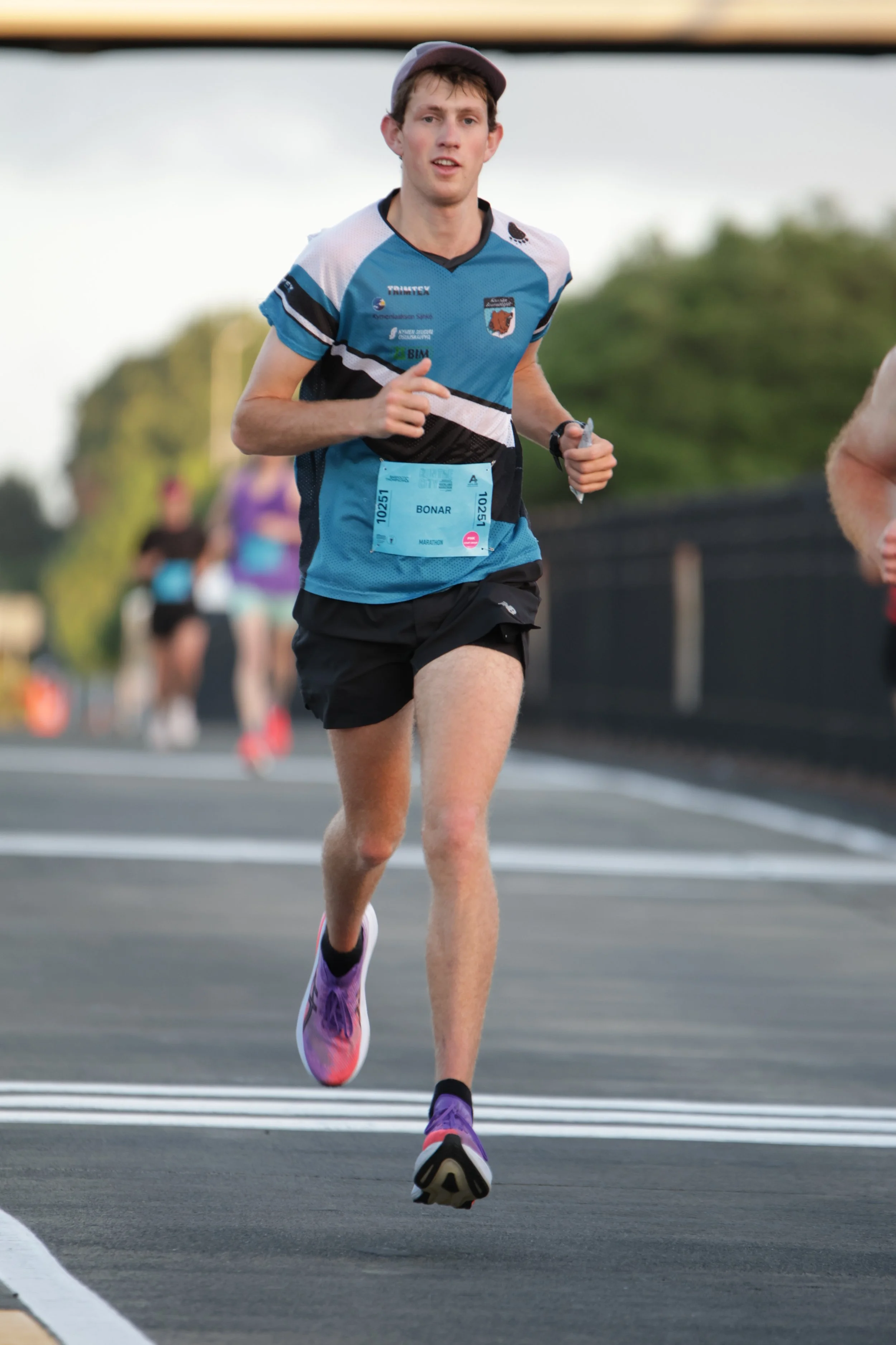 A male marathon runner running on a road wearing a blue and black athletic shirt, black shorts, purple running shoes, a grey cap, and a green bib with the name Bonar.