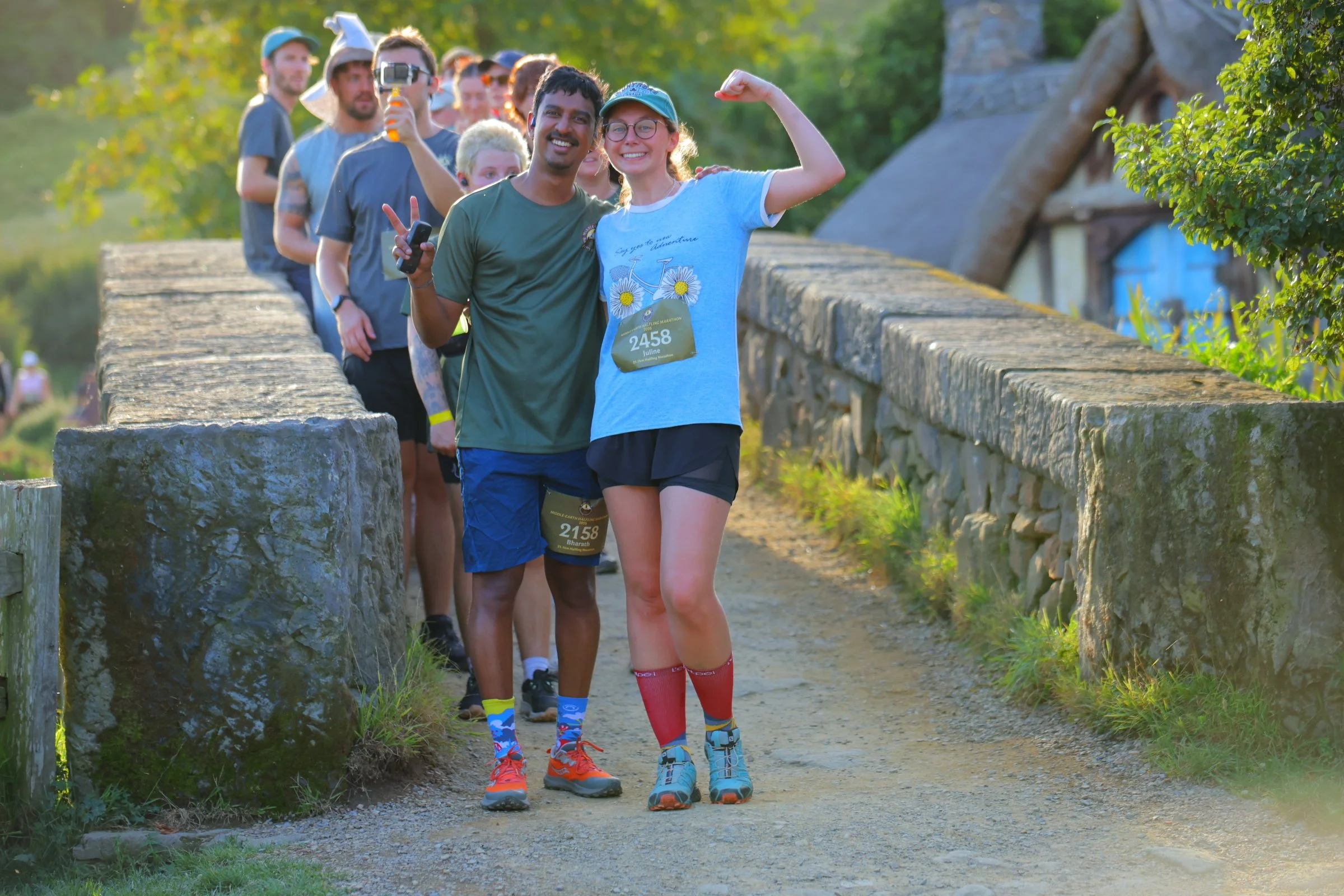 A group of marathon runners lining up on a rural path, with two women in the front flexing their muscles and smiling. They are dressed in athletic gear and wearing numbered bibs. The background features green trees and a stone wall along the trail.