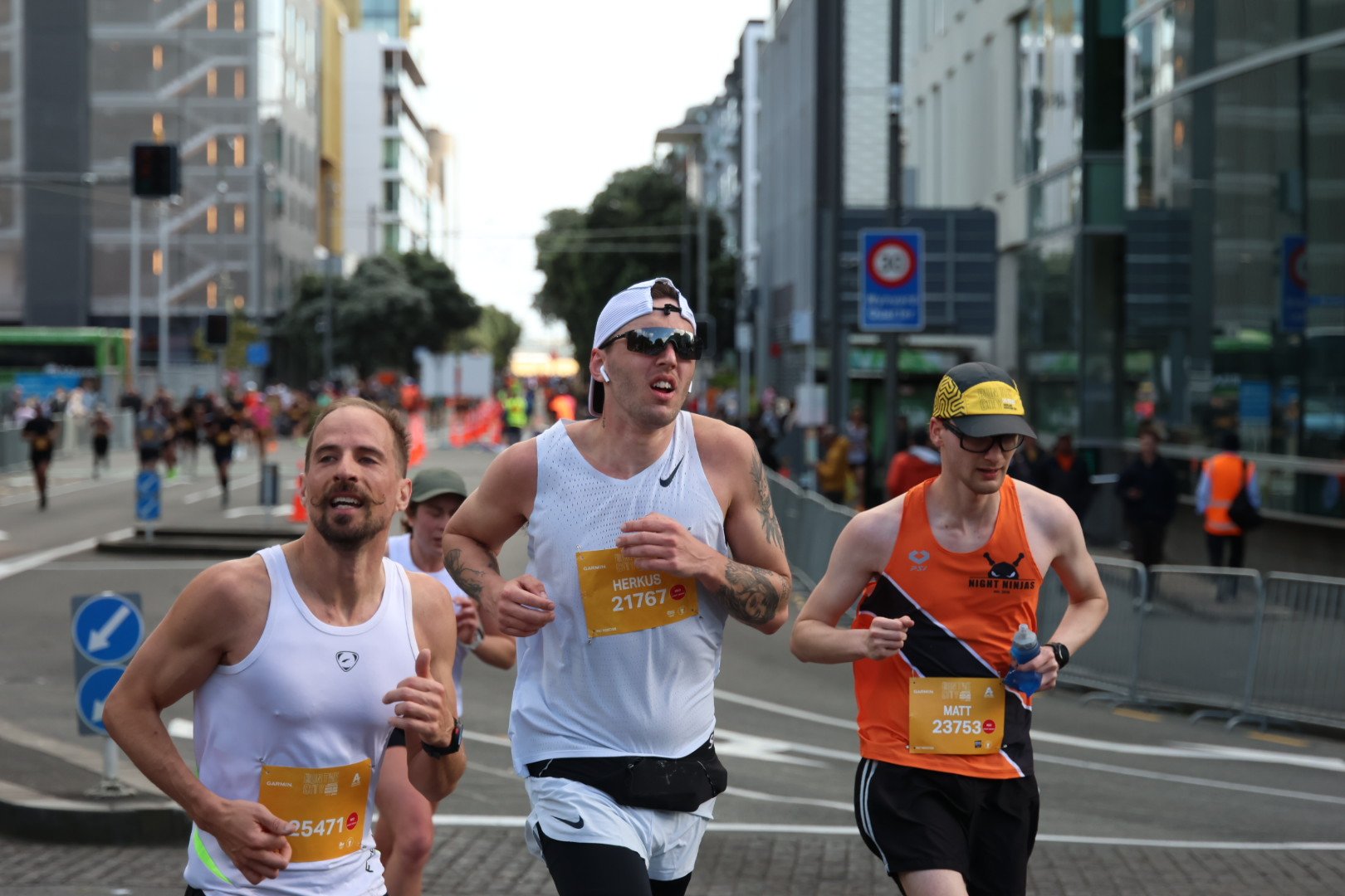 Three male runners participating in a marathon on a city street, wearing athletic attire and bib numbers, with other runners and city buildings in the background.