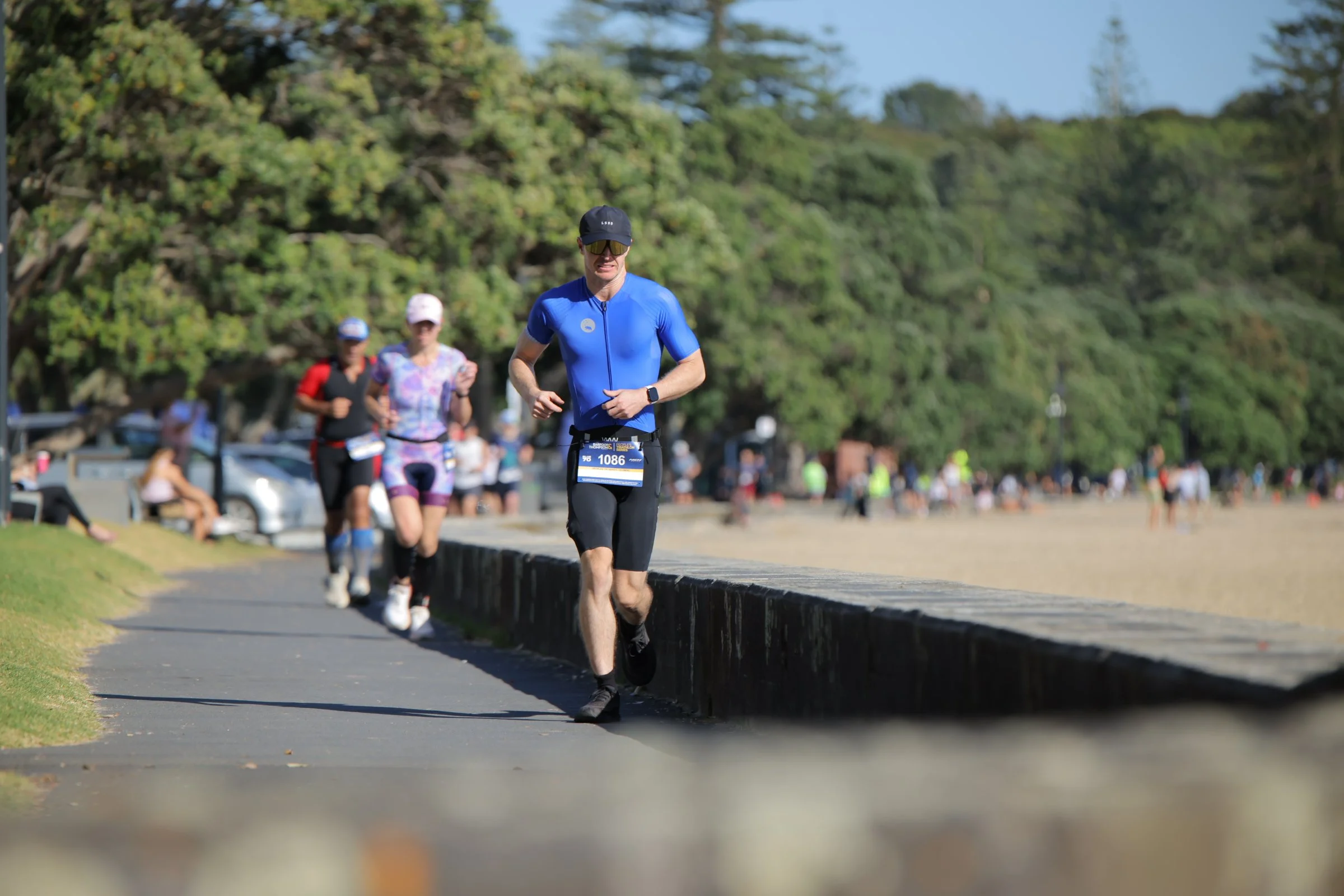 A group of runners participating in a marathon or race, running along a grassy park pathway with trees in the background.