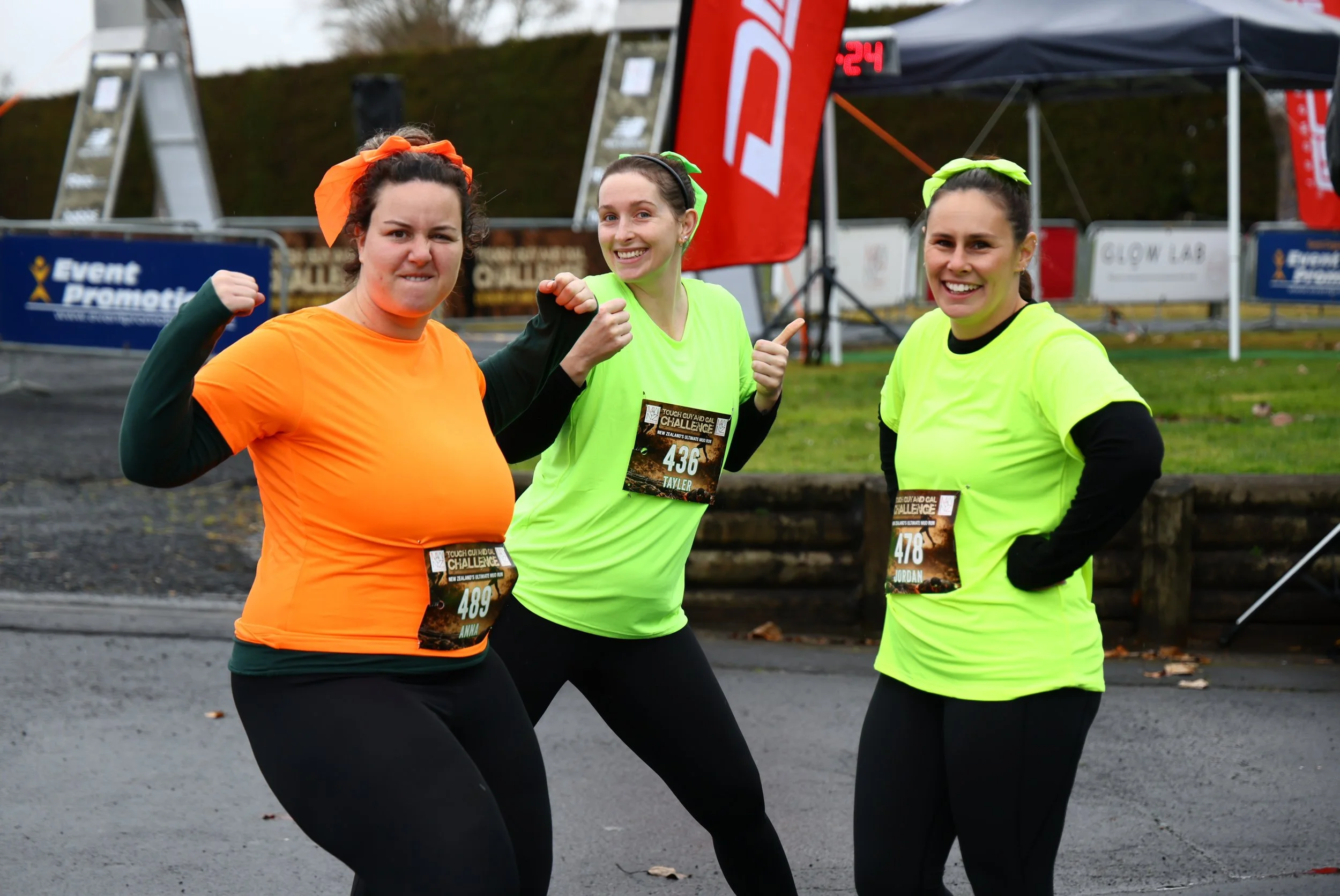 Three women in brightly colored running shirts pose for a photo after a race event, smiling and showing excitement.