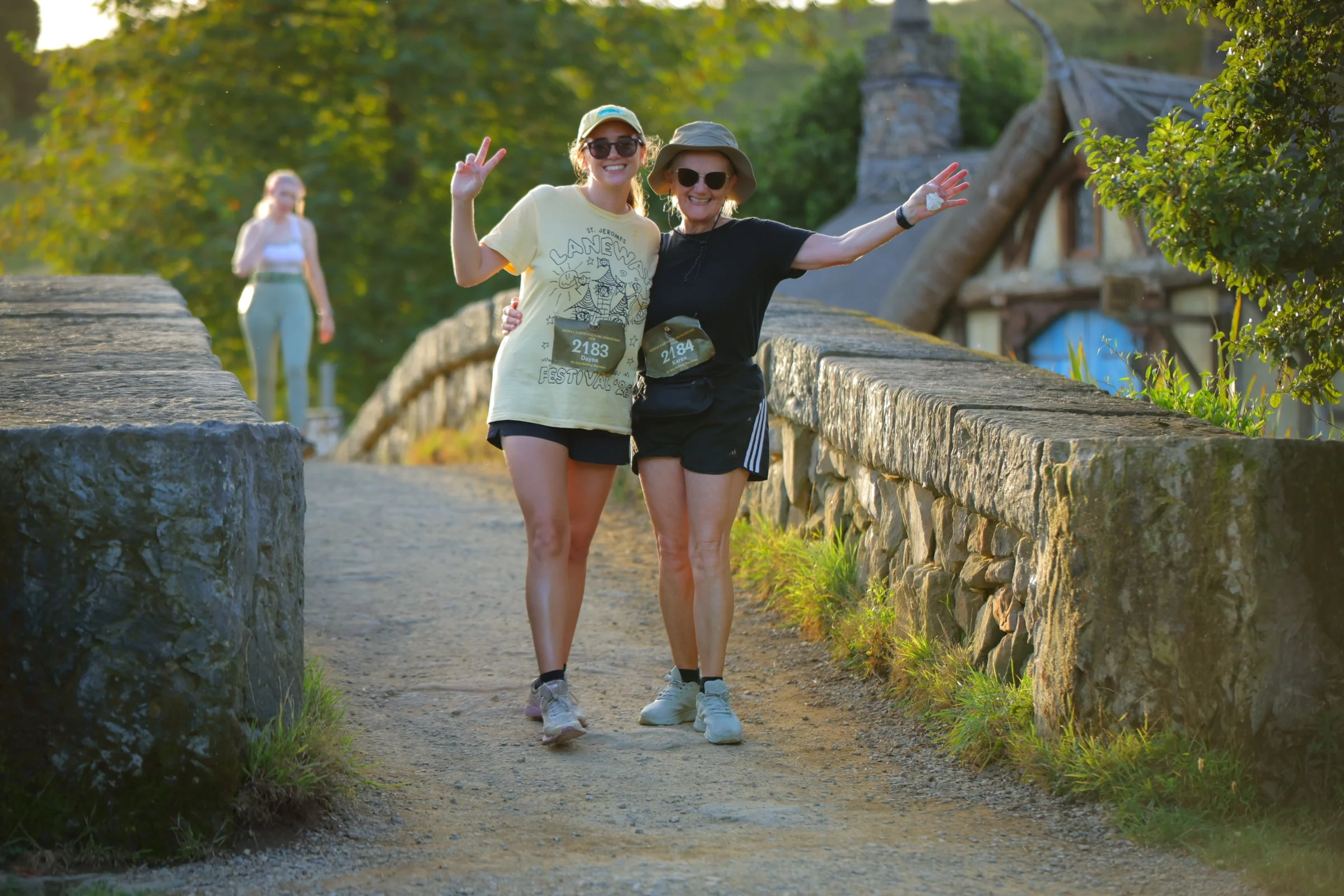 Two women smiling and posing on a stone bridge in a park during late afternoon or early evening, with one woman wearing a yellow T-shirt and the other in black, both wearing hats and sunglasses. In the background, a woman is walking along the bridge,