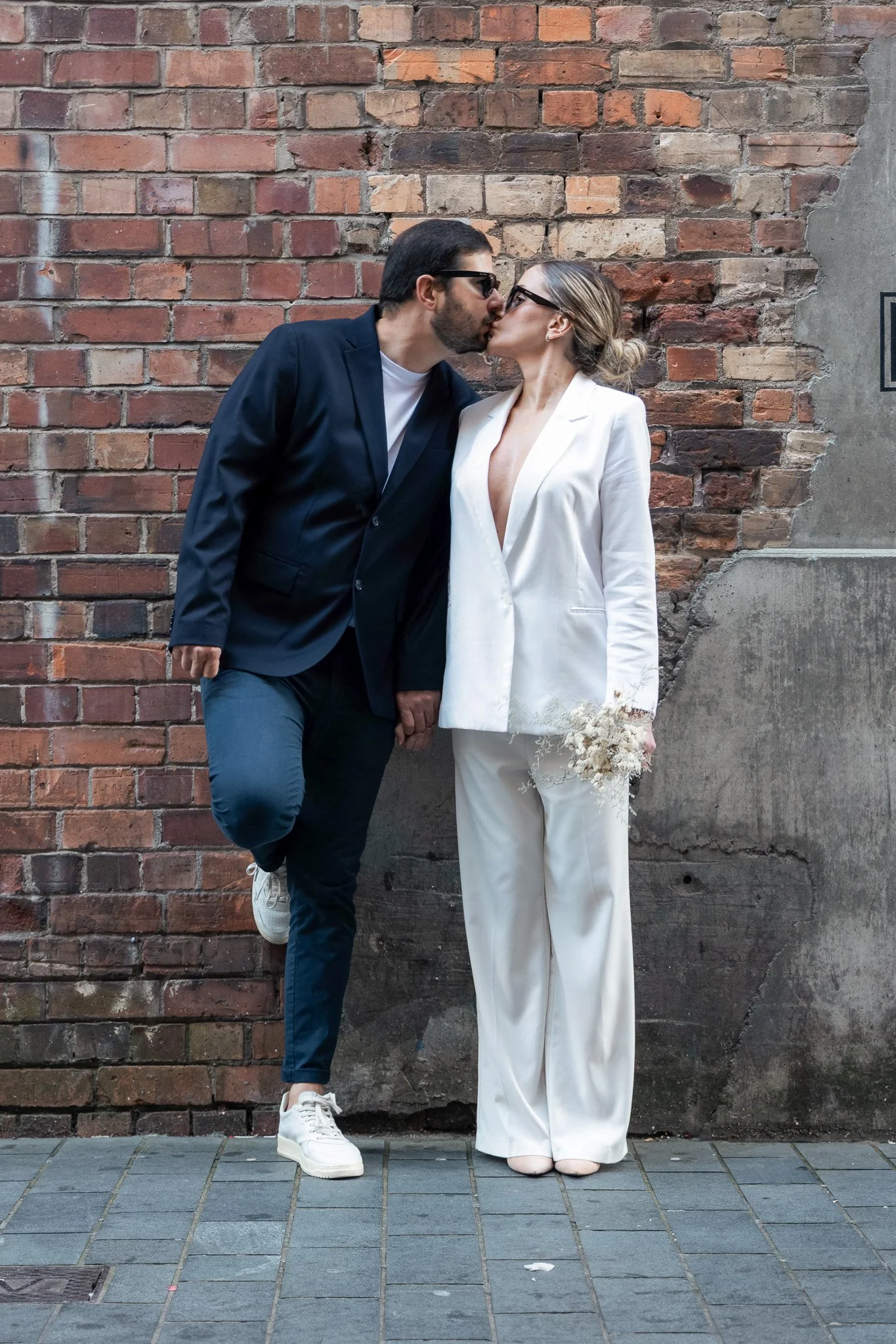 A couple kissing in front of a brick wall. The man is wearing a navy suit, white T-shirt, and sneakers, and the woman is dressed in a white suit and heels, holding a small bouquet. They are standing close, with the man on one leg, touching their lips