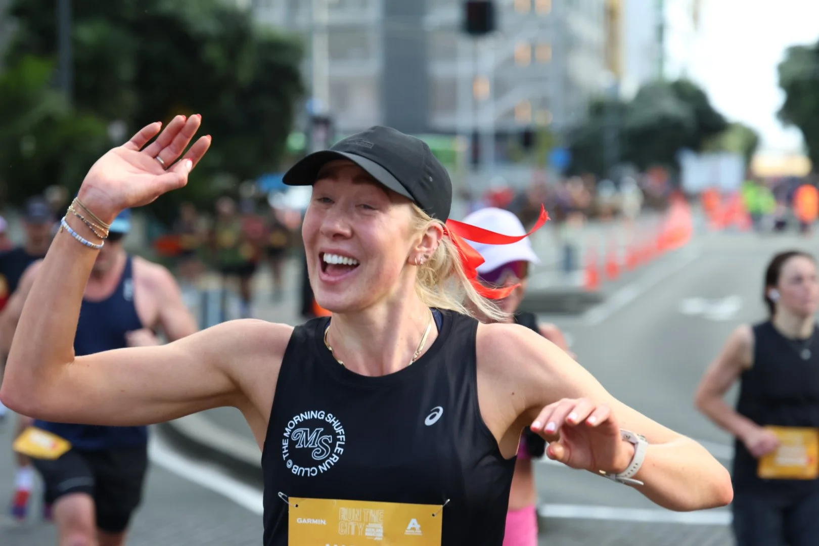 A woman smiling and waving during a marathon, wearing a black hat, black athletic tank top, and a yellow race bib, with other runners and orange traffic cones in the background.