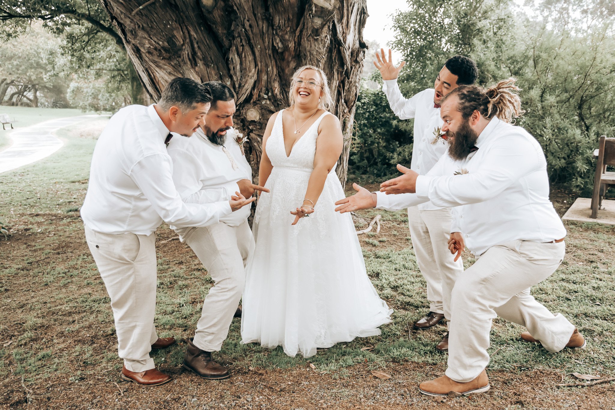 A bride in a white wedding dress and glasses standing under a large tree, surrounded by five men in white shirts and beige pants, who are playfully kneeling and gesturing towards her, outdoors in a park-like setting.