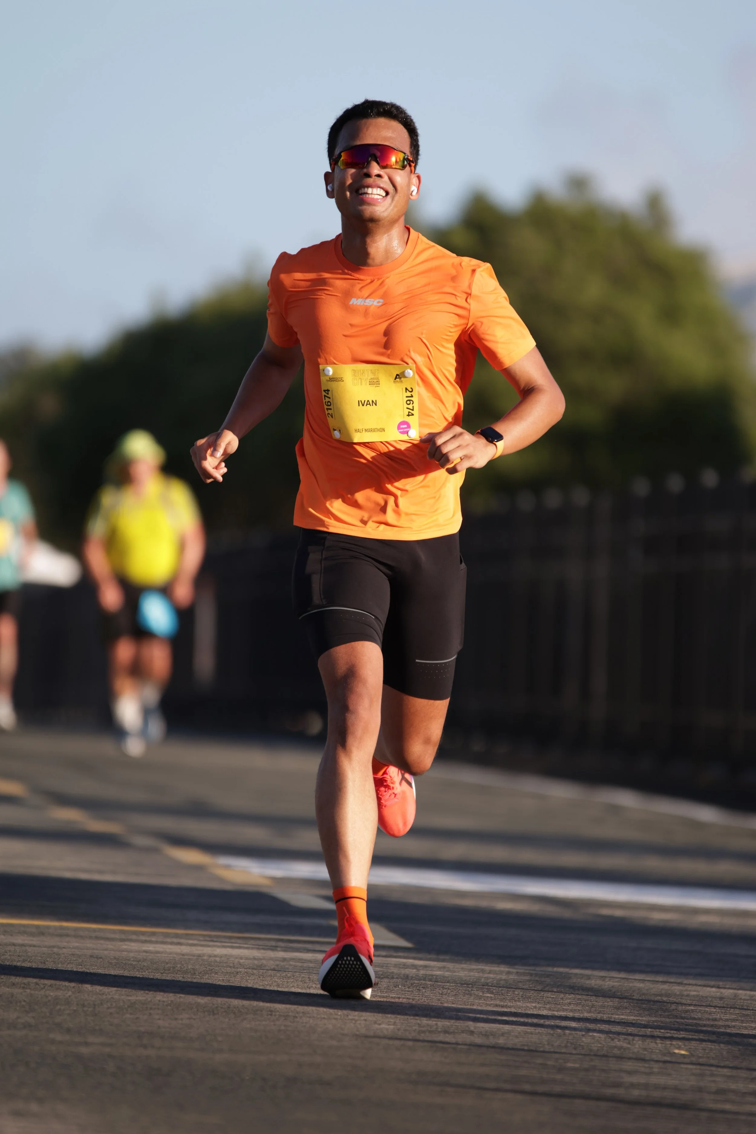 A man running in a marathon race, wearing an orange shirt, black shorts, orange shoes, sunglasses, a watch, and a bib with the name 'IVAN'. He is smiling and appears to be enjoying the run, with blurred runners and trees in the background.
