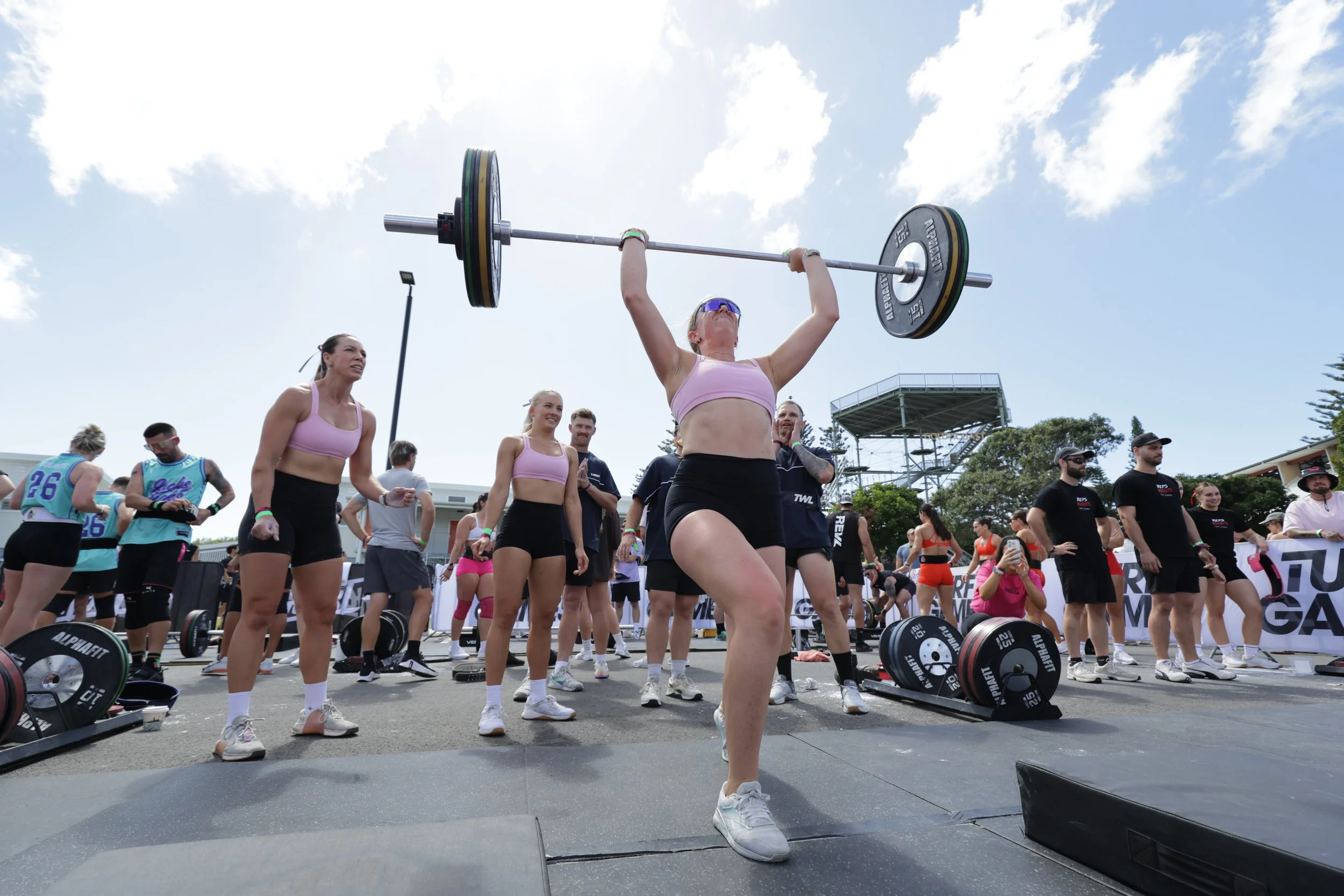 A woman in athletic attire preparing for a weightlifting event at an outdoor competition, surrounded by other athletes and spectators.