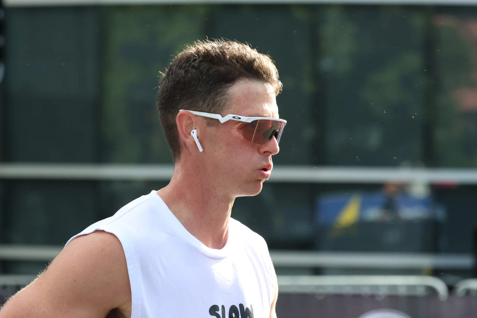 Profile of a young man with short brown hair, wearing sports sunglasses, wireless earbuds, and a sleeveless white shirt, outdoors with blurred background.