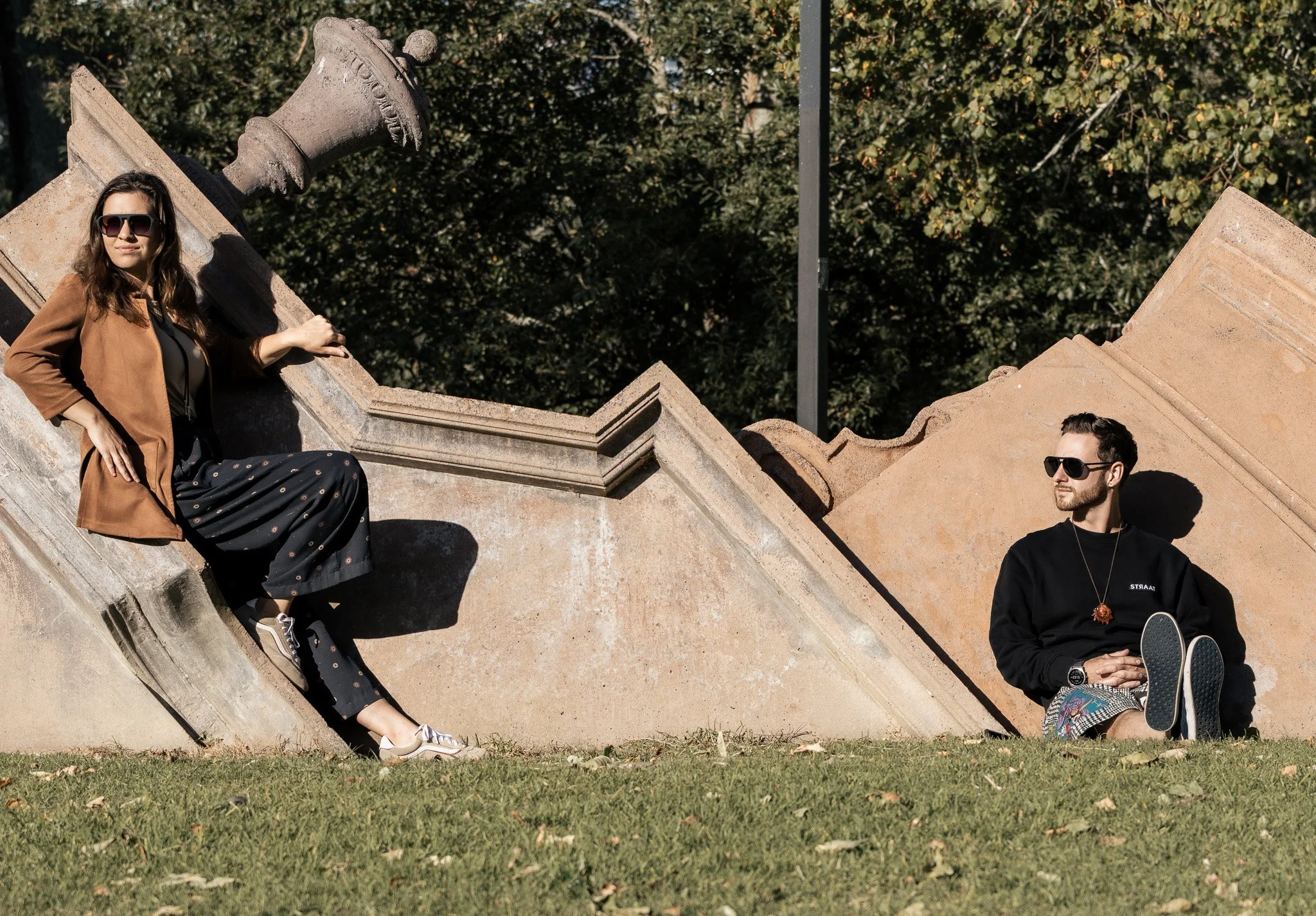 Two young adults sitting on a stone monument in a park with trees in the background. The woman is wearing sunglasses and a brown coat, and the man is wearing sunglasses and a black sweatshirt. They are relaxing outdoors.