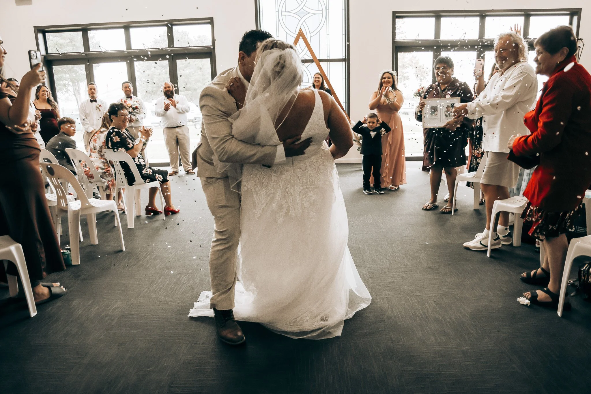 A newlywed couple sharing a kiss during their wedding ceremony in a bright indoor venue, surrounded by family and friends celebrating and throwing confetti.