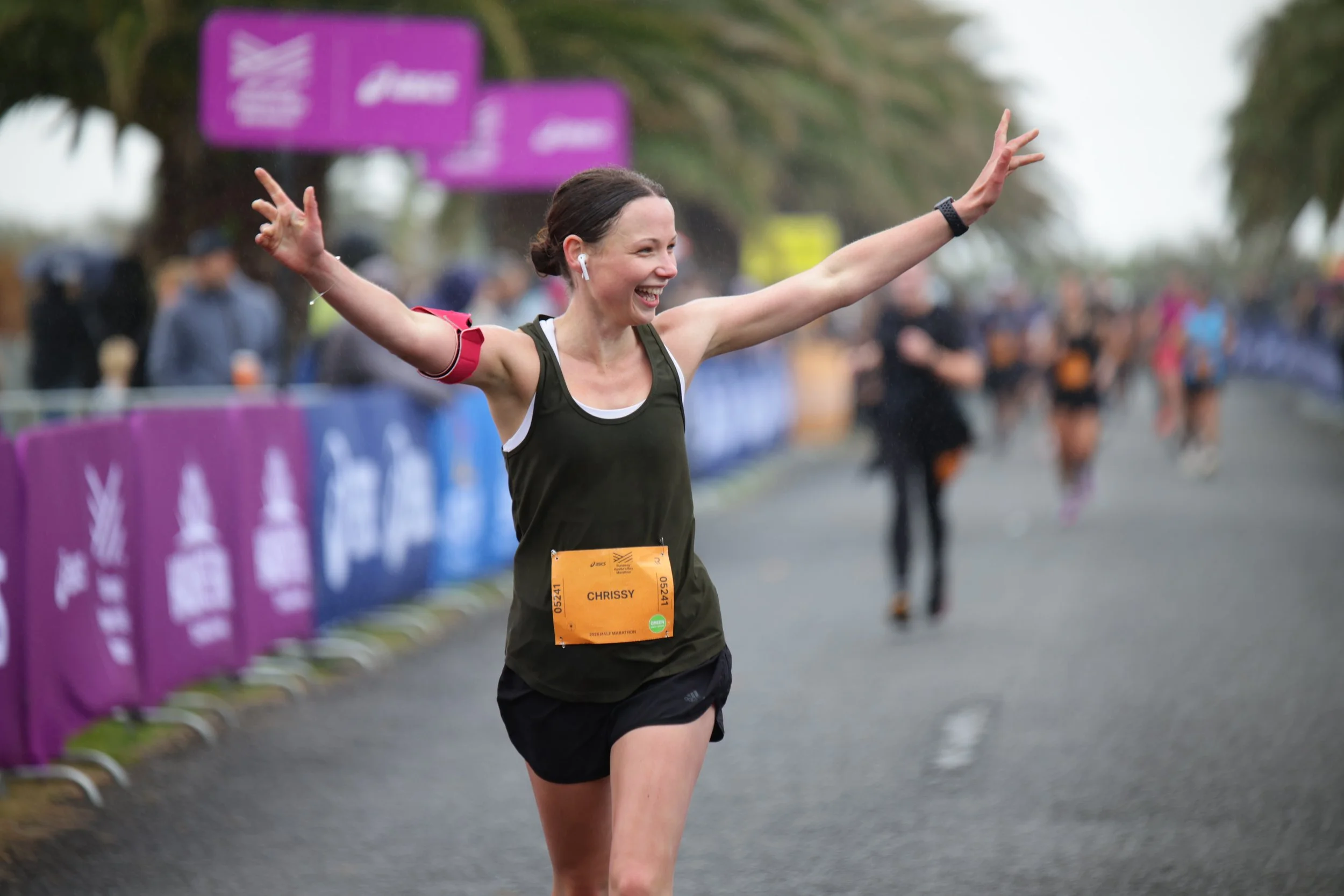 A female marathon runner crossing the finish line with arms raised, wearing a black tank top and shorts, with a race bib that says 'CHRISSY'. She is smiling and wearing wireless earbuds. The background shows a blurry crowd and purple banners along th
