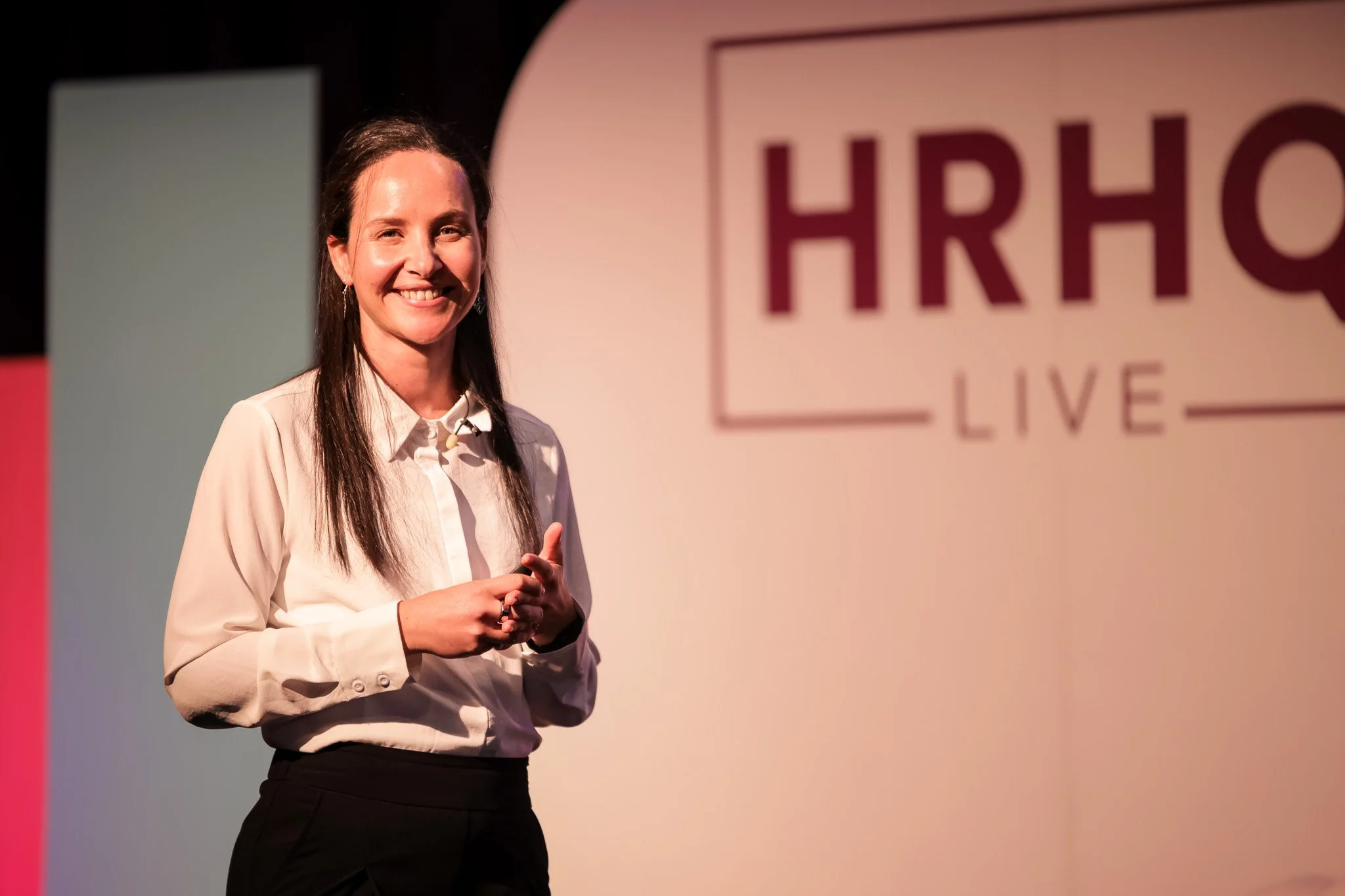 A woman with long dark hair smiling and speaking on stage at an event with a large sign in the background that reads 'HRO LIVE'.