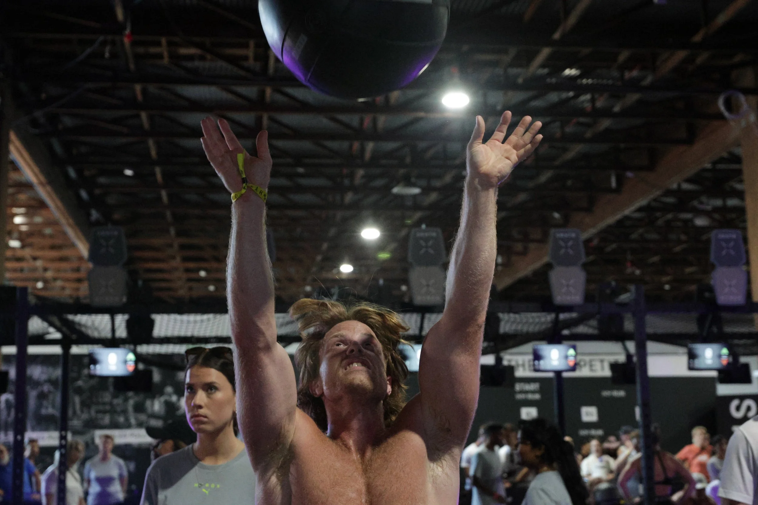 A person with long hair and a yellow wristband is jumping and reaching toward a black ball hanging from the ceiling of an indoor gym or sports venue, with other people in the background.