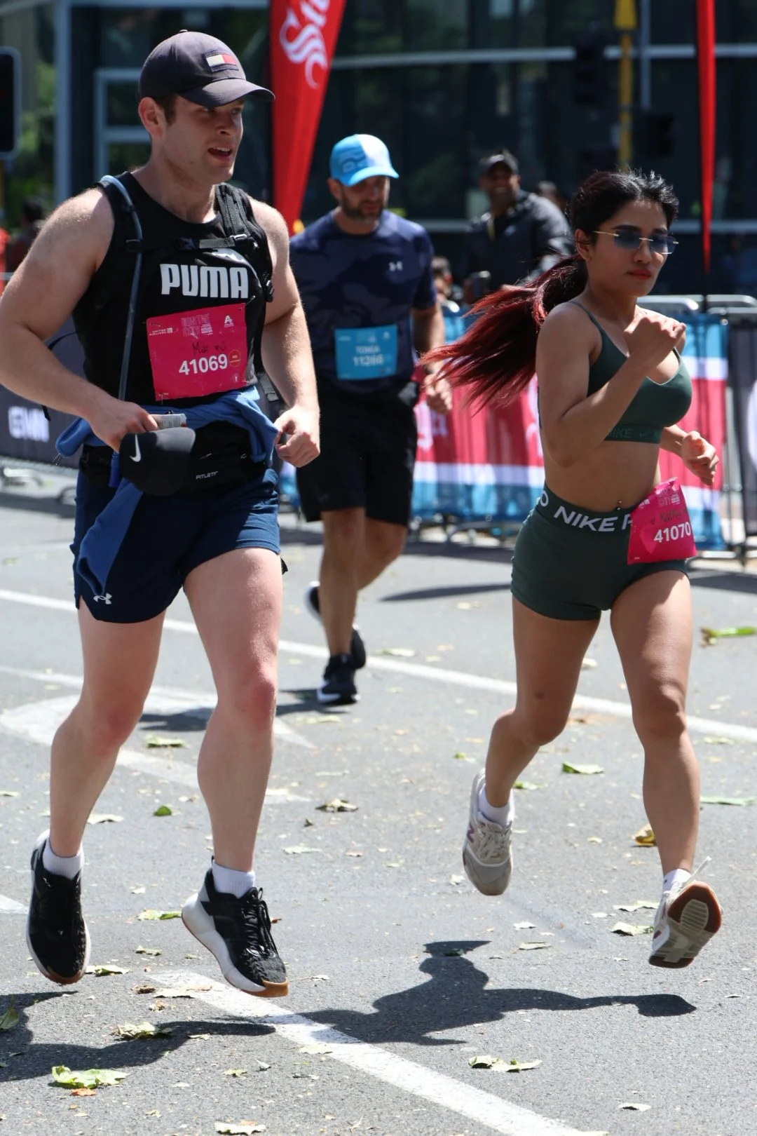 Two runners, a man and a woman, running in a marathon on a city street on a sunny day, with background spectators, banners, and fallen leaves on the ground.