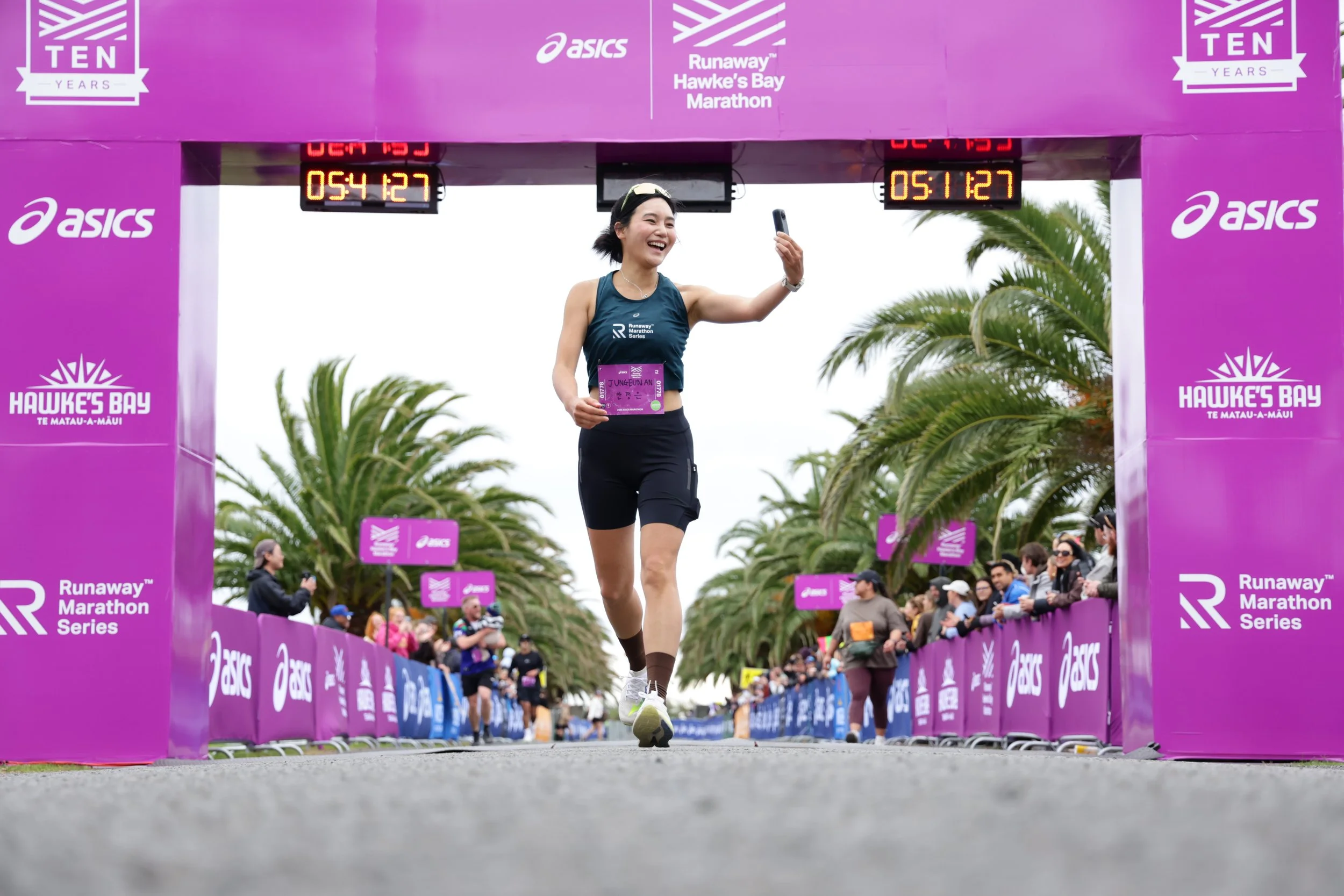 A female runner crossing the finish line at the Hawke's Bay Marathon, smiling and taking a selfie, with timer and race banners visible.