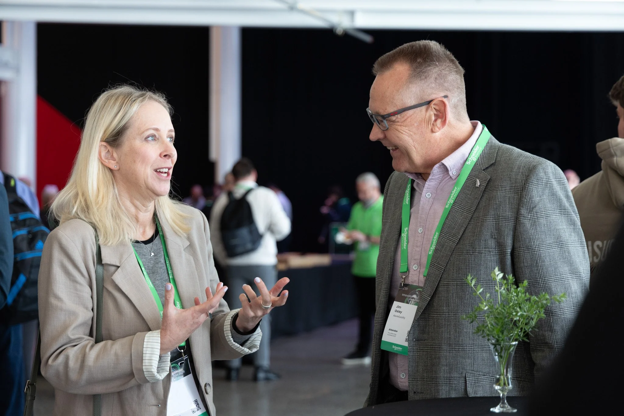 A woman and a man are engaged in conversation at a conference, wearing name badges on green lanyards. The woman has long blonde hair and is gesturing with her hands, while the man, wearing glasses and a checkered blazer, is smiling. There are other a
