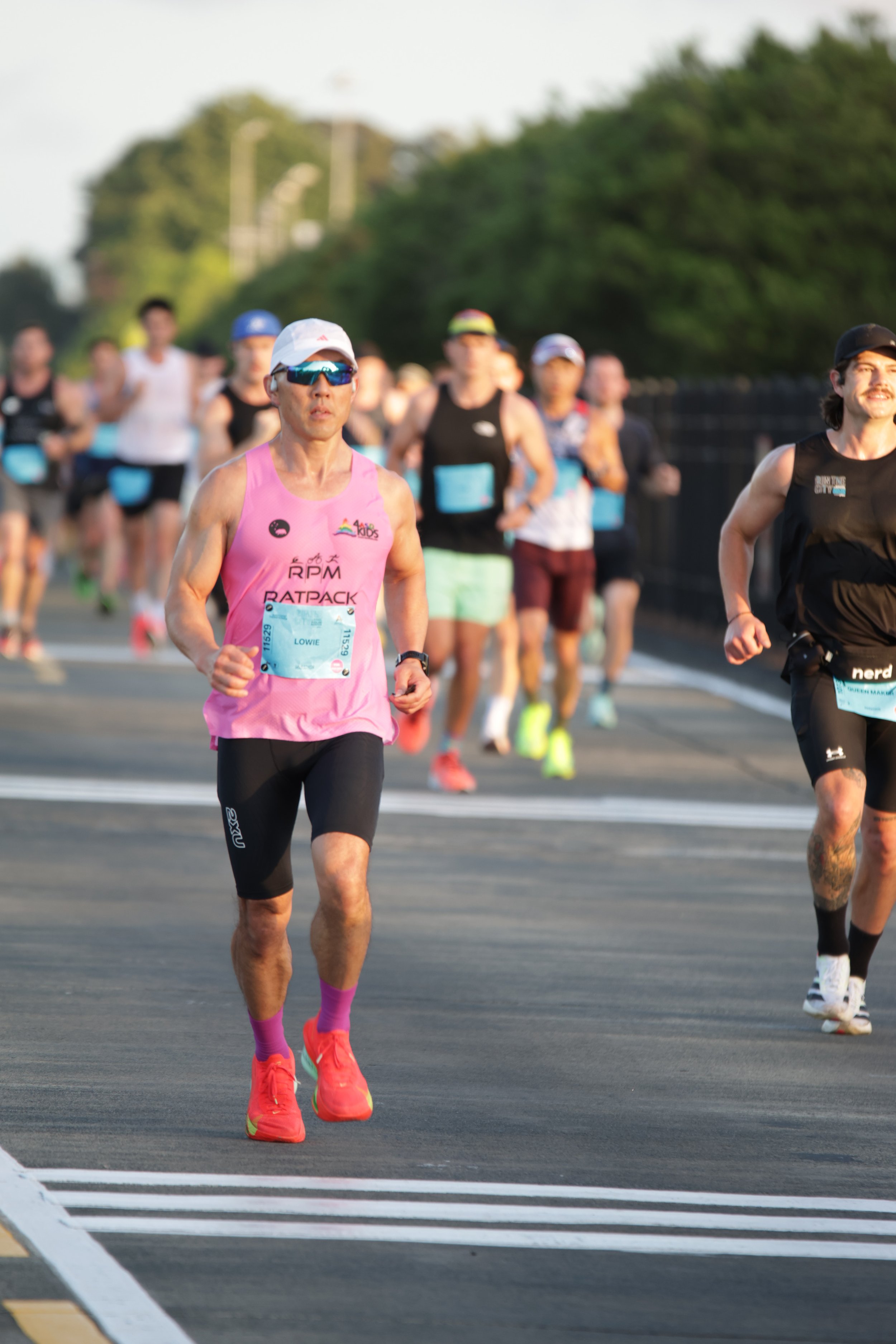 Male marathon runner wearing pink tank top, black shorts, pink running shoes, sunglasses, and a white cap, running on a road with other runners in the background during a race.