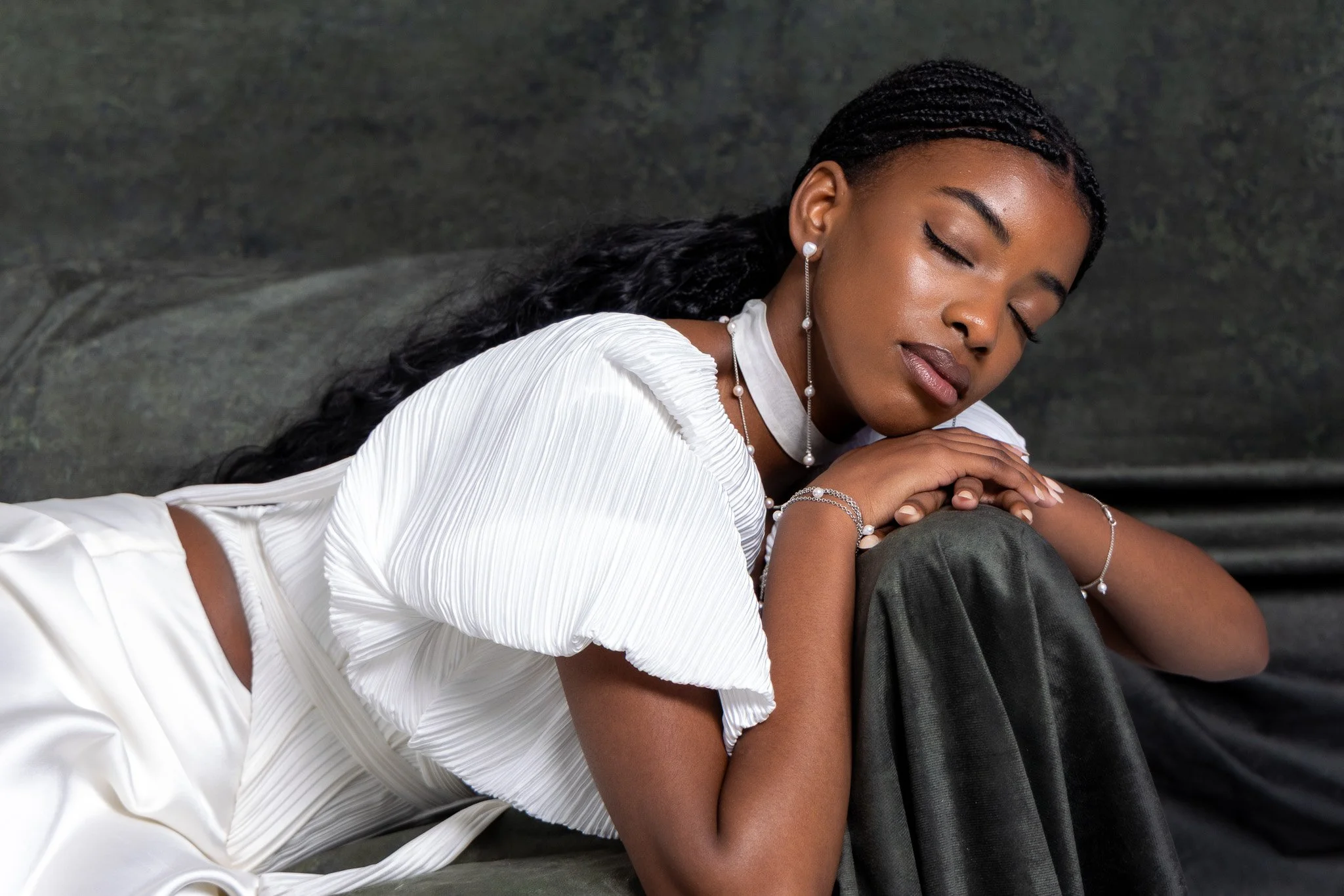 A young woman with dark braided hair wearing pearl jewelry and a white pleated dress, sitting with her eyes closed and her arms resting on her knees, against a dark textured background.
