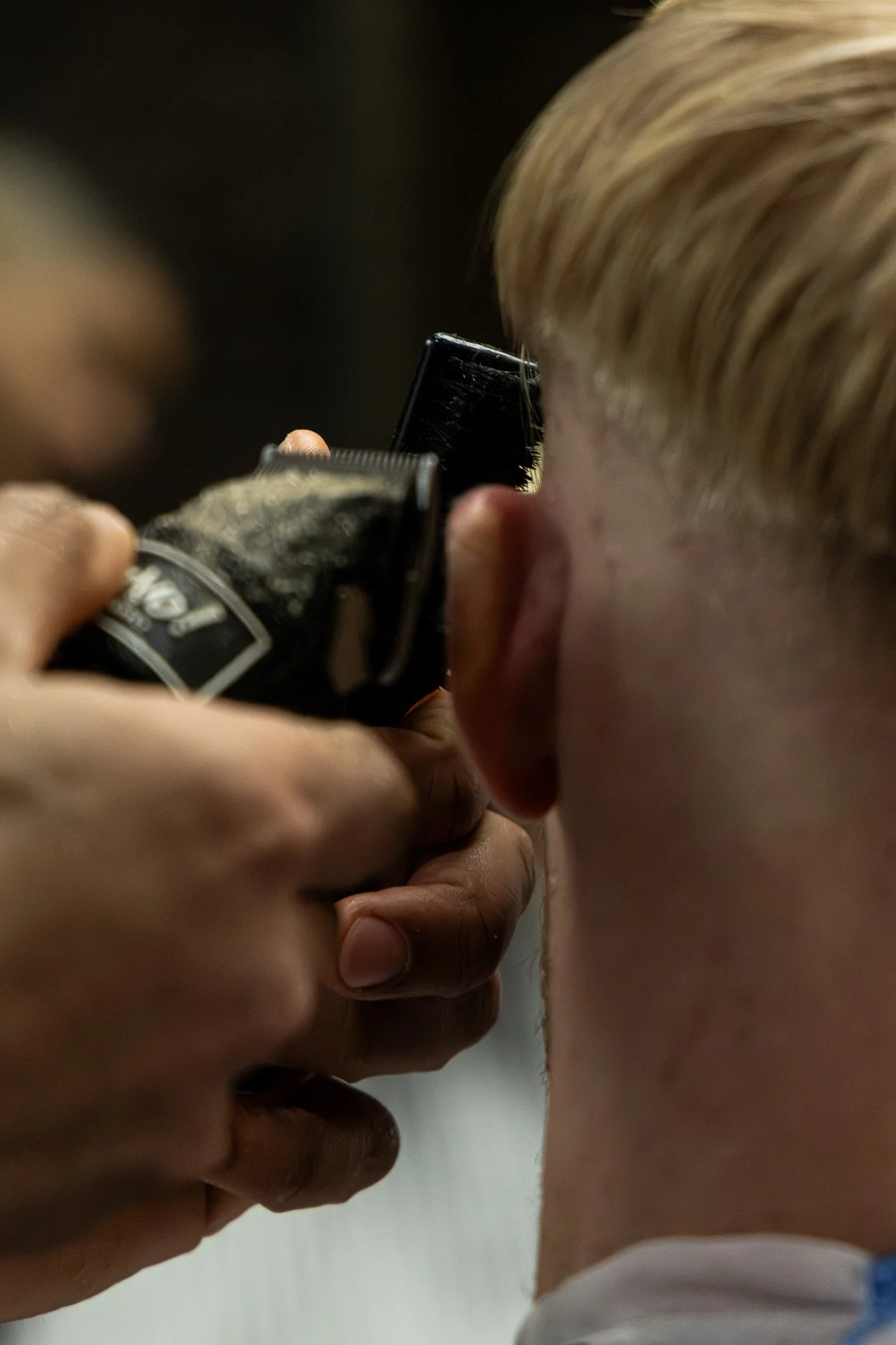 A person getting a haircut with an electric clipper, focusing on the side of their head and hairdresser's hands.