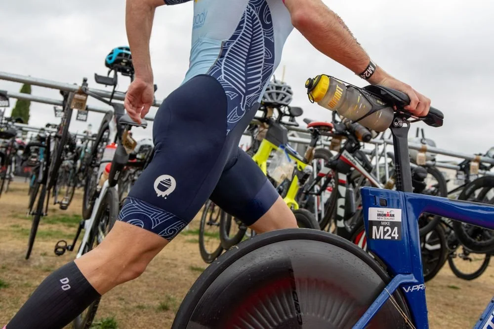 Close-up of a cyclist's lower body and bike, with bicycles parked in the background at a race or event.