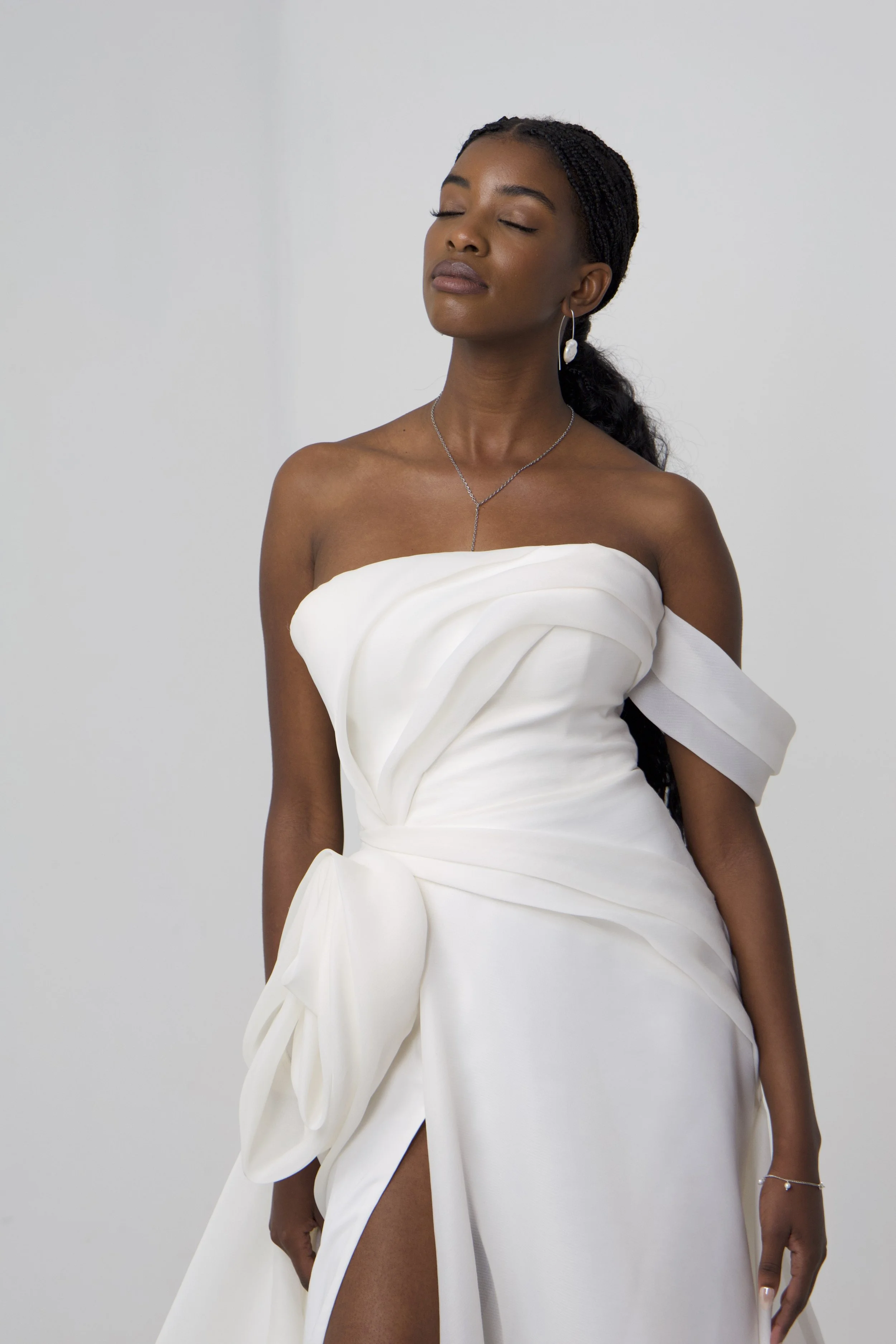 A woman in a white strapless gown with bow details on the side, standing with her eyes closed against a plain light background, wearing jewelry including earrings, a necklace, and a bracelet.