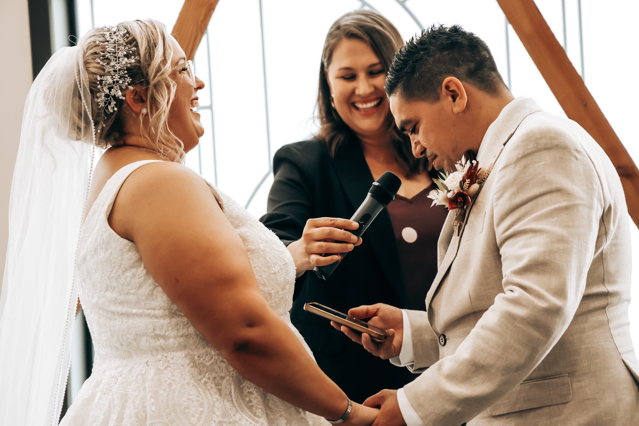 A wedding ceremony with a bride and groom holding hands and exchanging vows, accompanied by an officiant in front of a glass window.