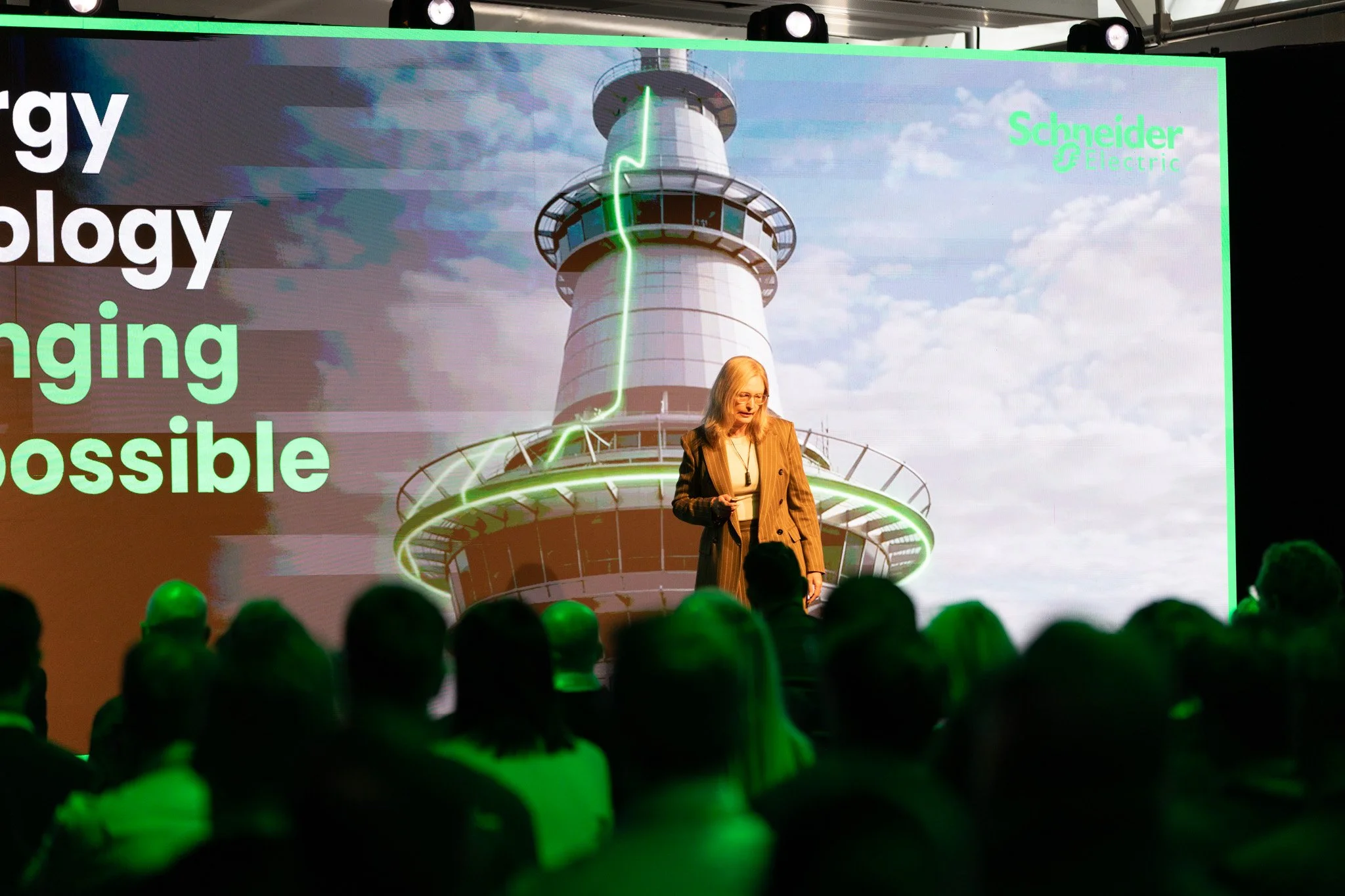 A woman with blonde hair and glasses giving a presentation on stage in front of an audience. Behind her, a large screen displays an image of a tower with a green lightning bolt and the words 'energy,' 'ecology,' 'changing,' and 'possible.' The screen