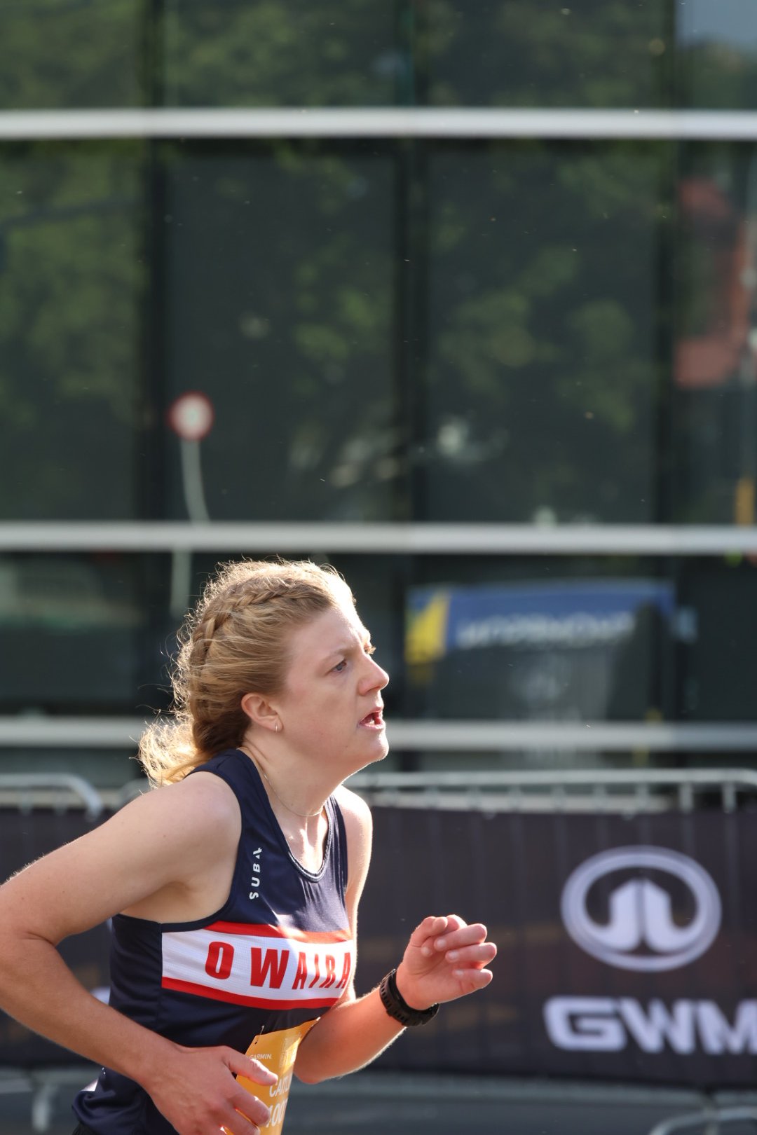 A woman running in a race, wearing a navy blue and red athletic uniform with the text 'O Wain' on it, and a yellow race bib. She appears focused and is outdoors.