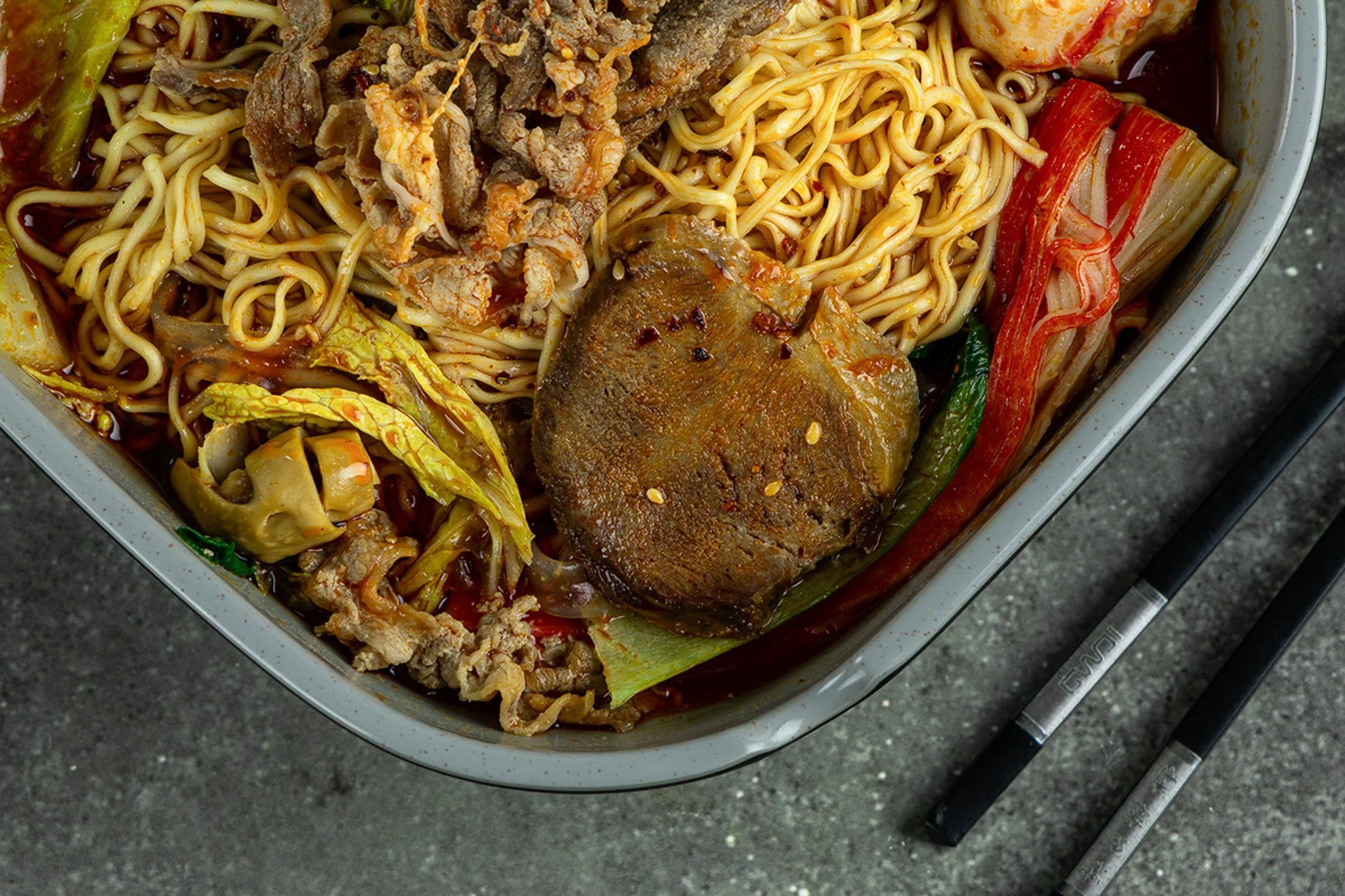 A close-up of a container filled with Asian-style noodle soup, featuring noodles, vegetables, a large meatball, and strips of beef or pork, topped with sauce.