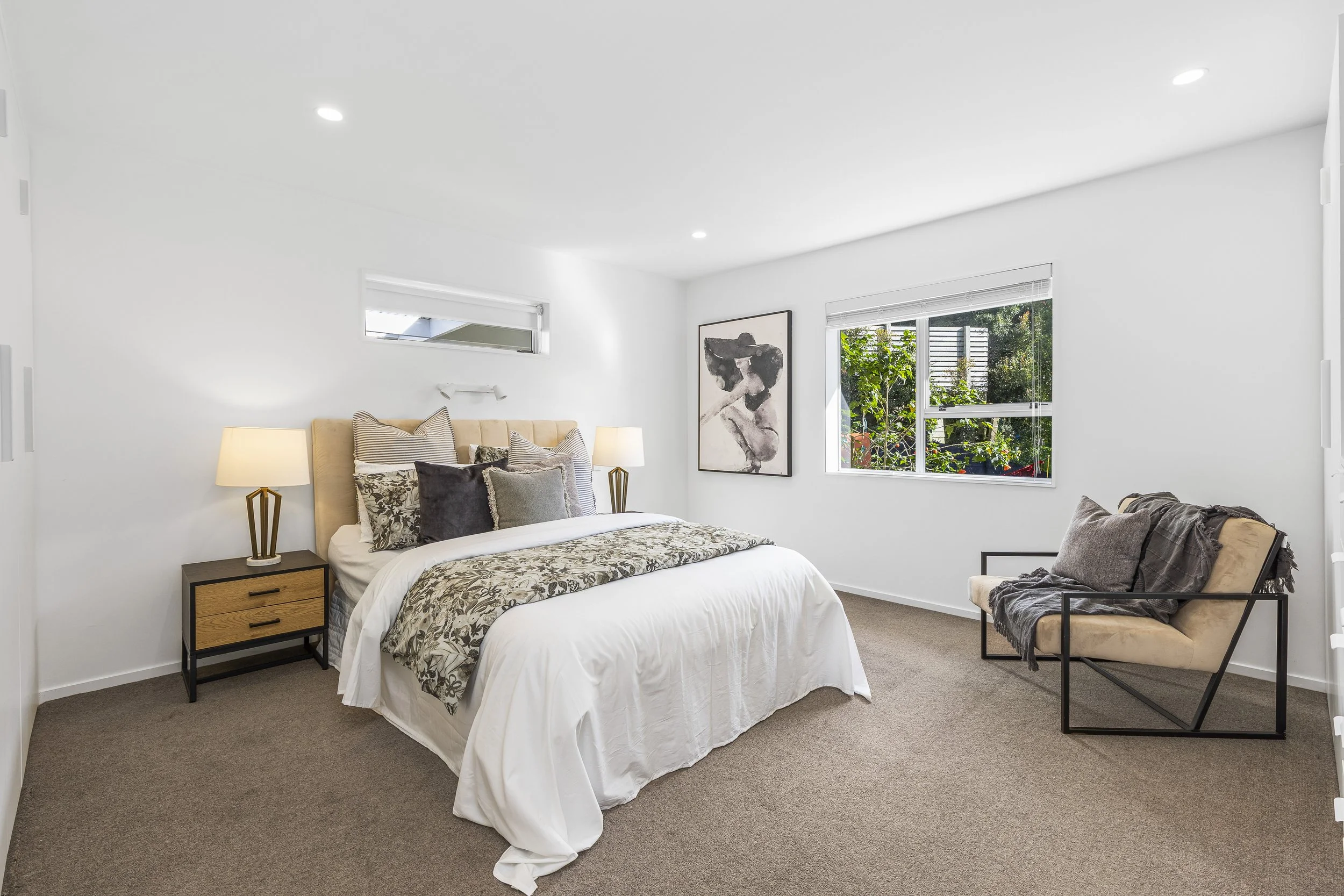 A neatly made bed in a bedroom with white walls, a beige headboard, and patterned and solid pillows, flanked by wooden nightstands with lamps. There are two windows—one small and high, another larger—letting in natural light and revealing greenery ou